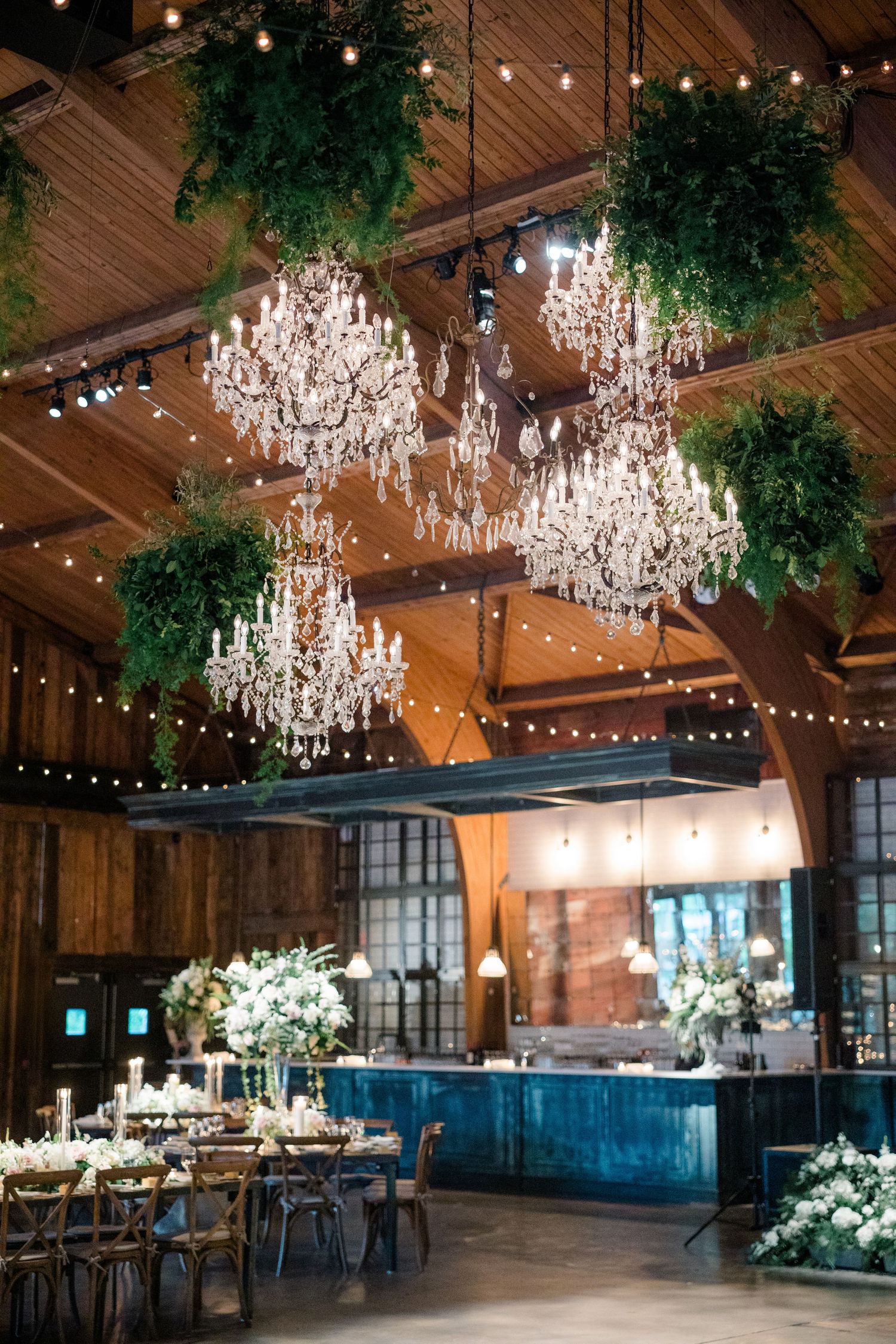 Elegant wedding reception venue with crystal chandeliers, hanging greenery, and wooden beams in rustic indoor space.