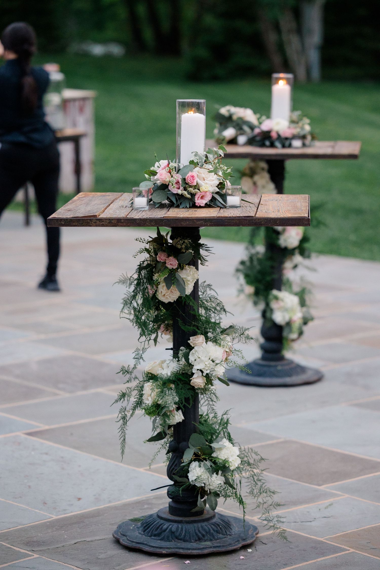 Decorative cocktail tables with floral garlands and candles on ornate metal pedestals at outdoor event.