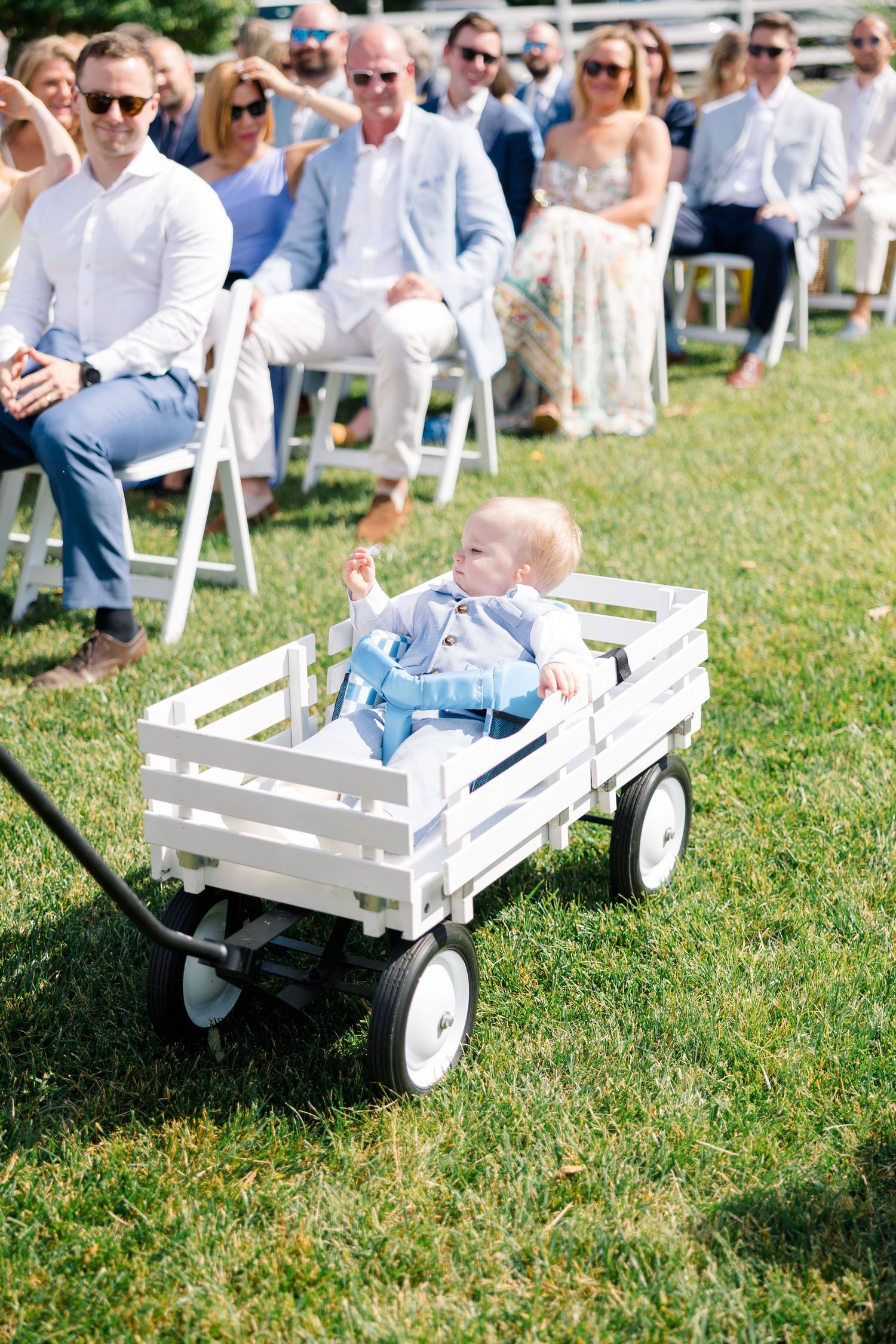 Adorable baby in blue outfit riding a white wooden wagon down the aisle at an outdoor wedding ceremony.
