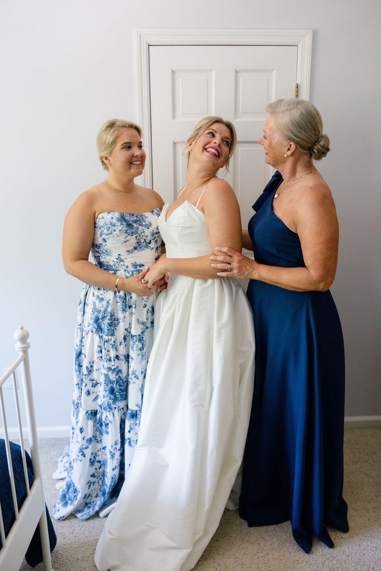 A bride in a white gown shares a joyful moment with two women in blue dresses during wedding preparations.
