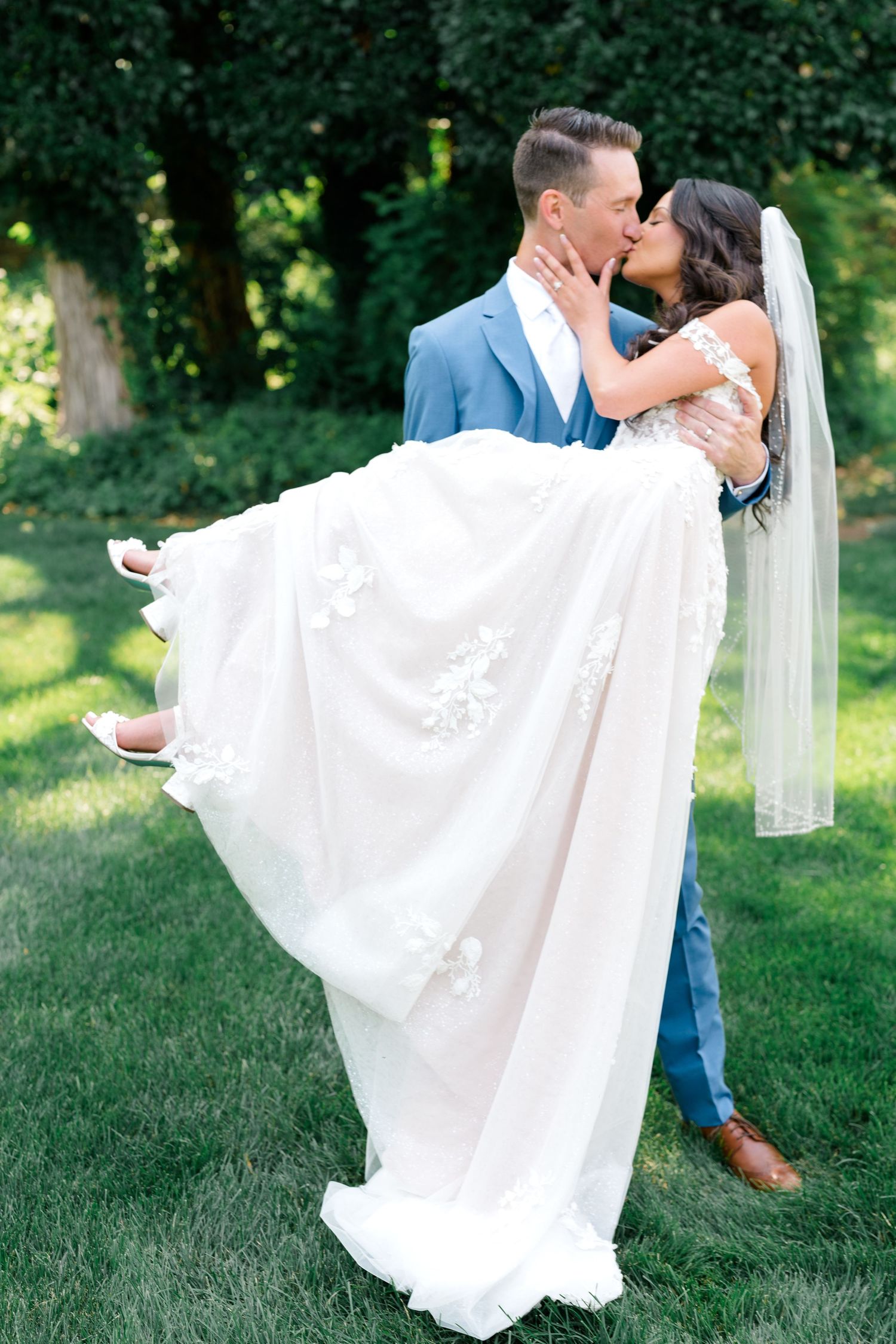 Groom lifts bride in flowing white dress during romantic outdoor wedding photo.