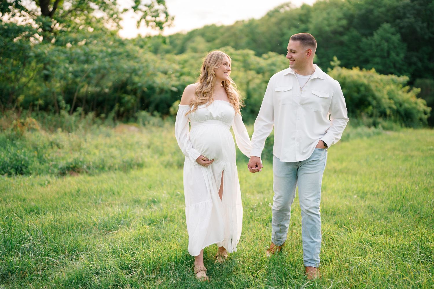 Romantic maternity shoot in green meadow with couple wearing coordinated white and denim outfits.