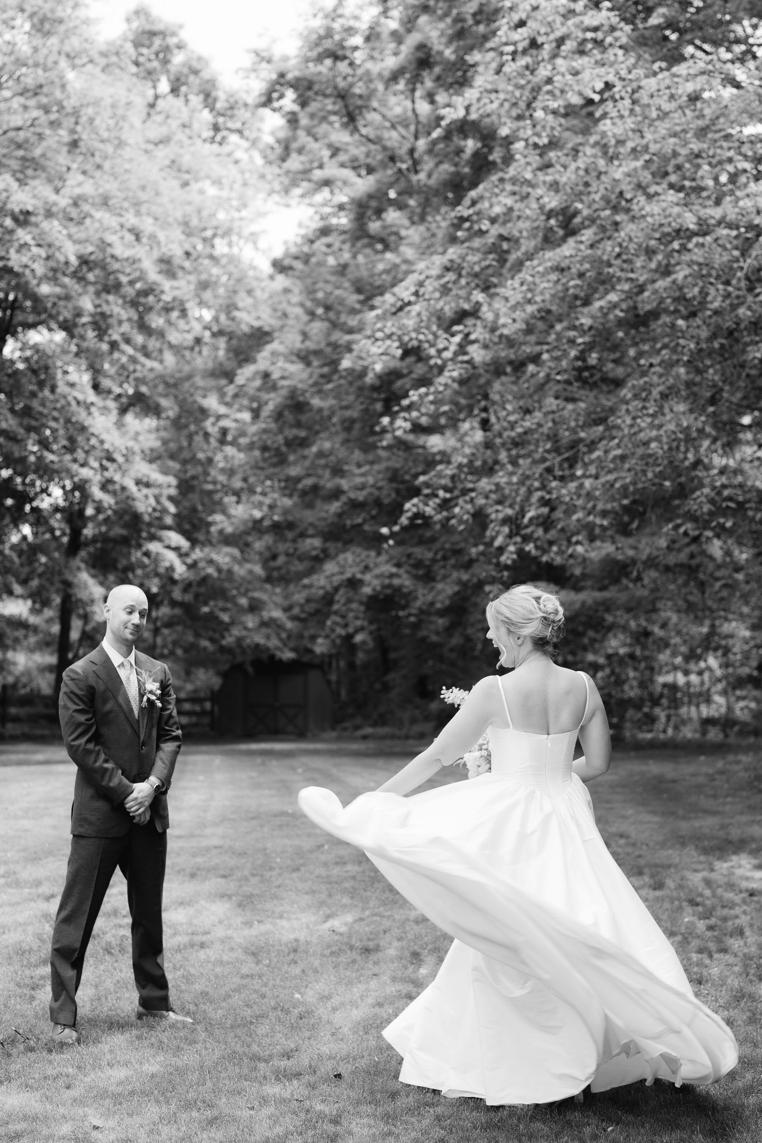 Black and white photo of groom watching bride twirl in flowing wedding gown during first look outdoors.
