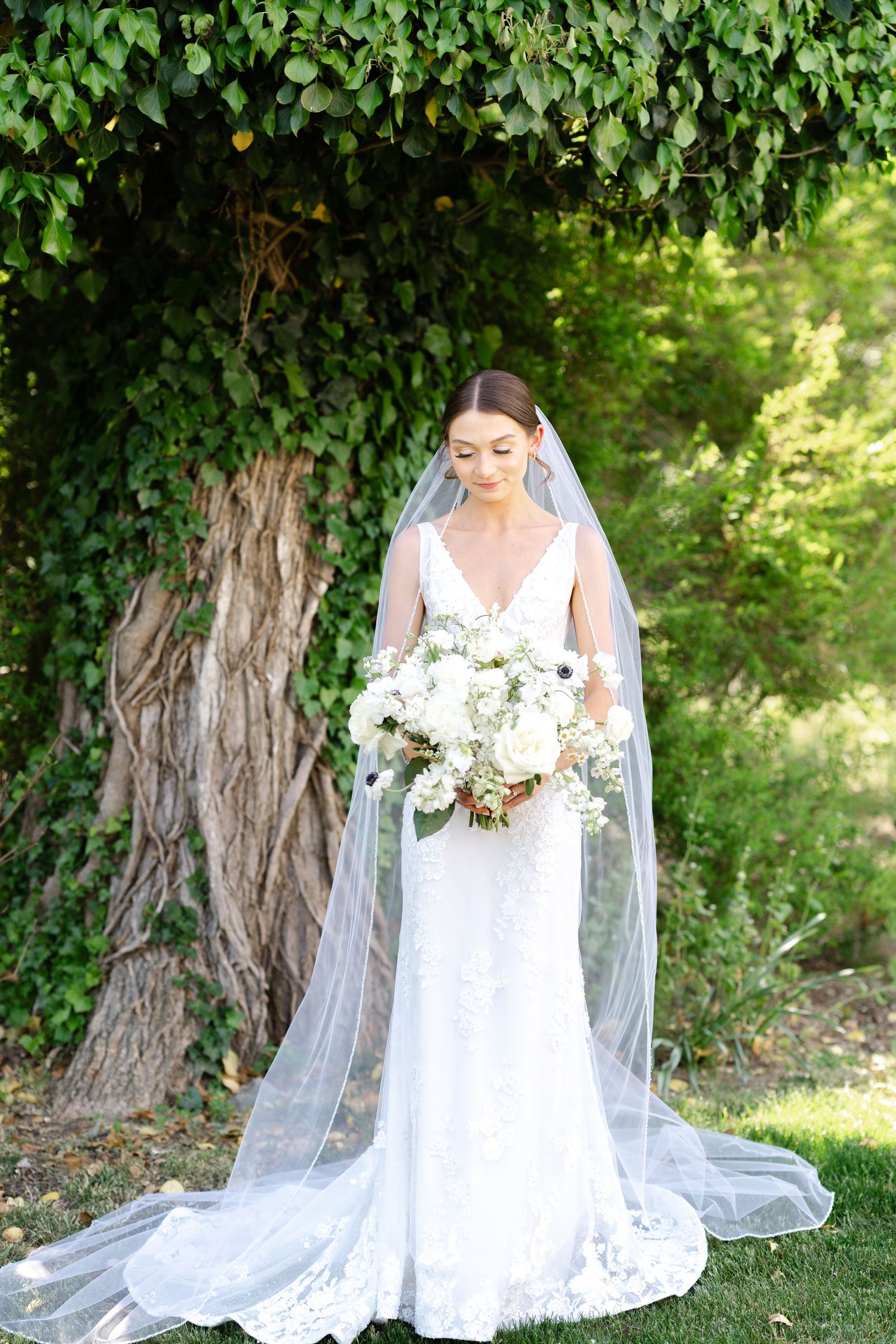 A bride wearing a flowing white lace gown and veil poses on grass near trees during an outdoor photo session.