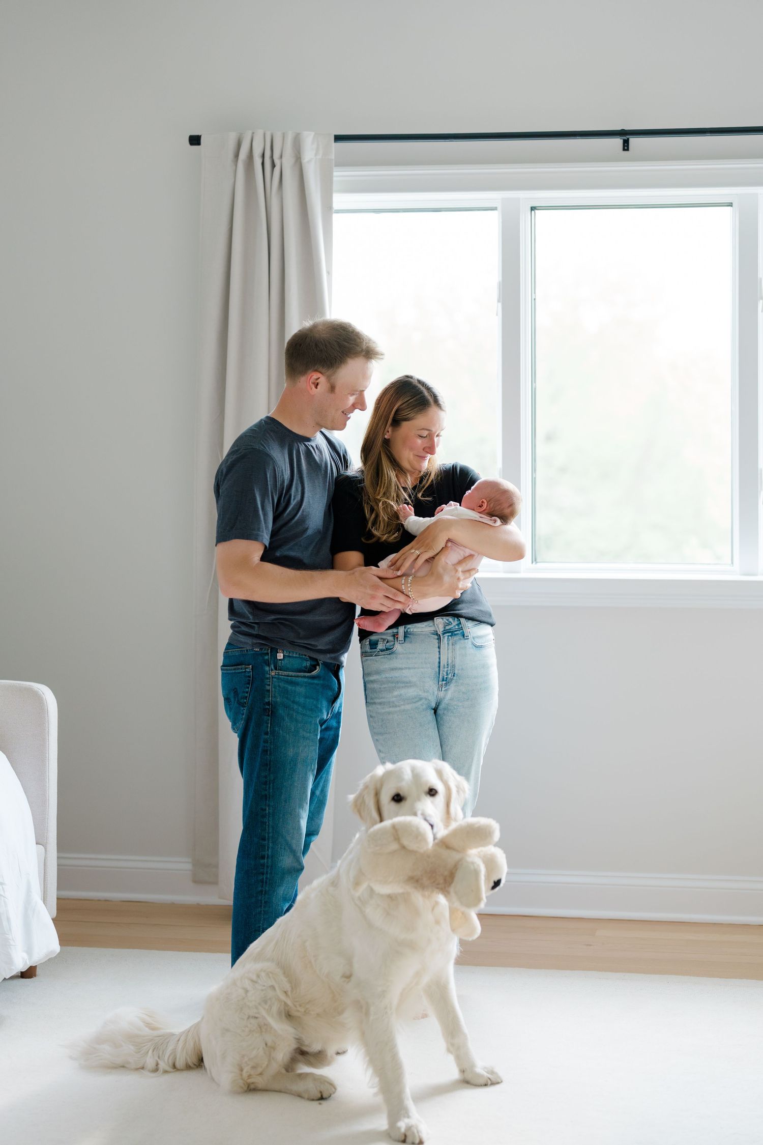 A couple stands by a window with their newborn baby while their golden retriever sits nearby.