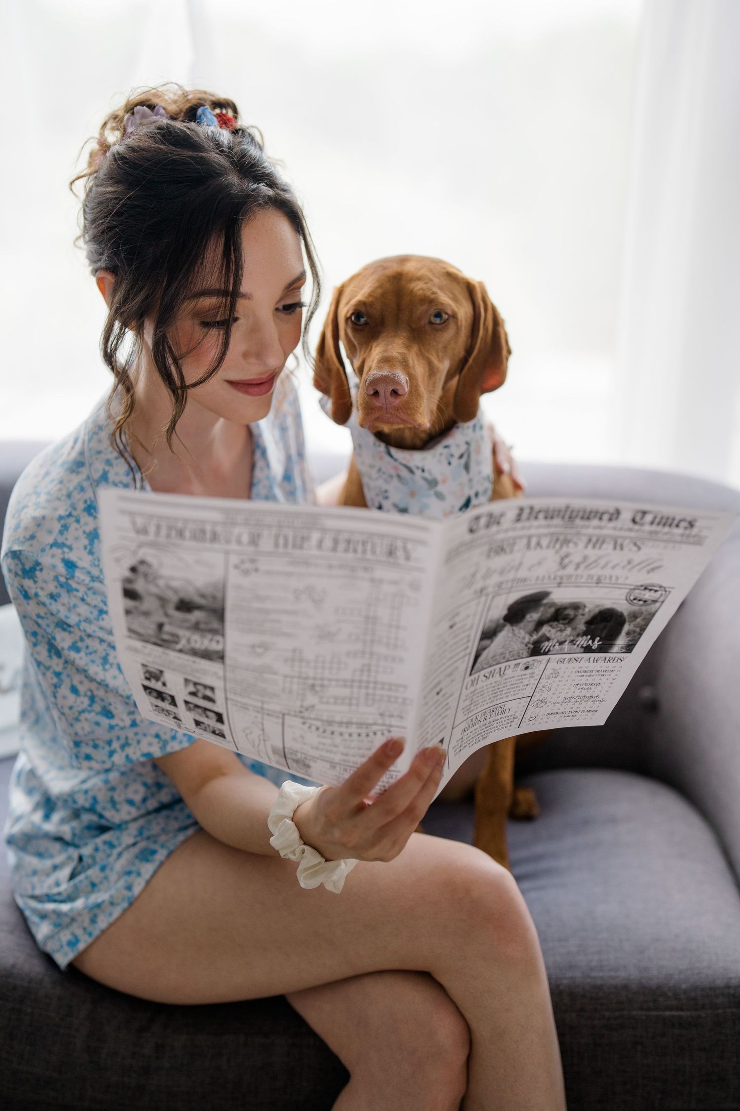 Woman in blue dress reads newspaper with attentive Vizsla dog on grey couch in bright room.