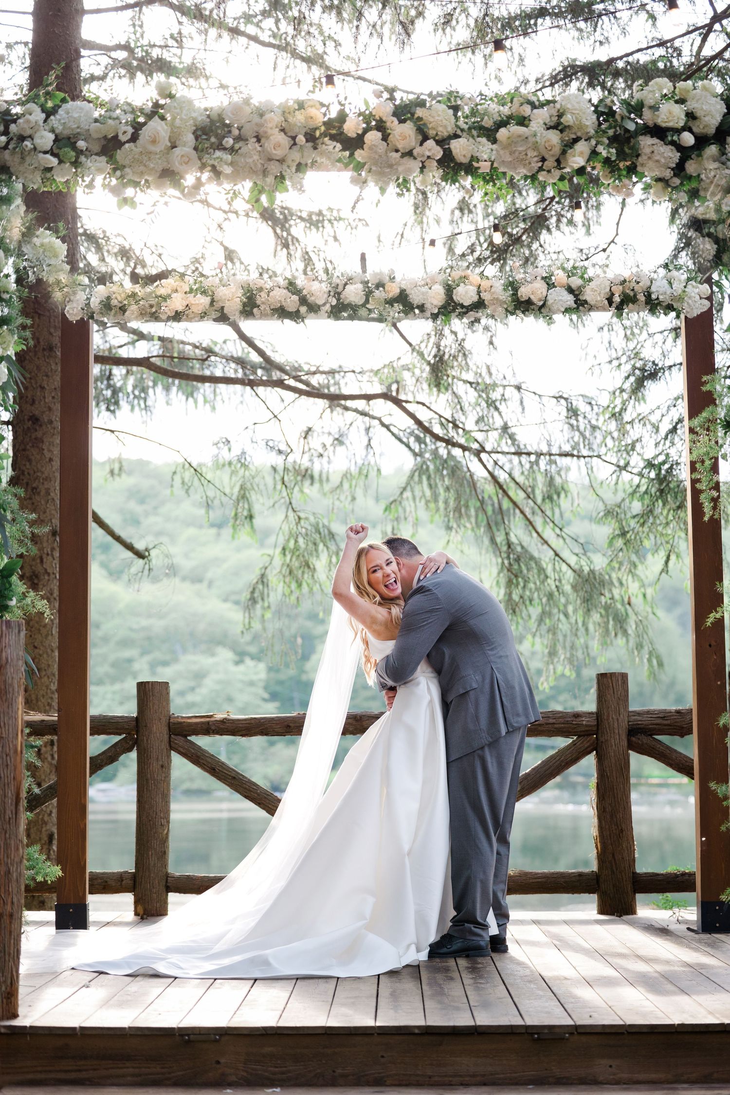 Romantic wedding moment on a deck overlooking a lake under an elegant white floral arch.
