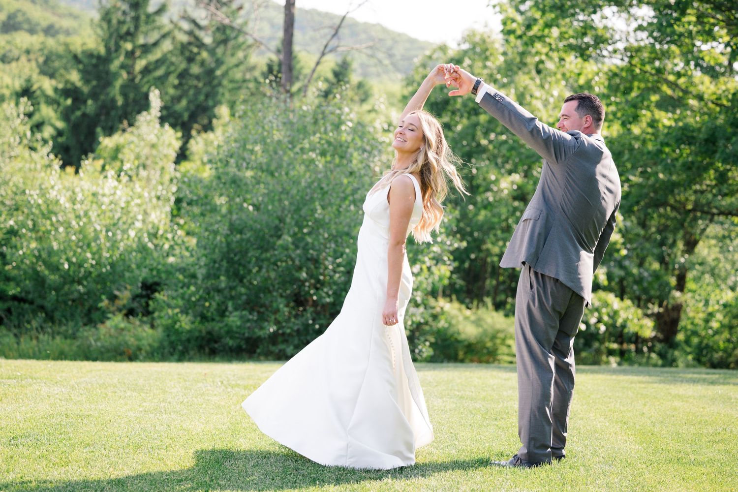 A playful wedding couple dancing and twirling on a grassy hillside.