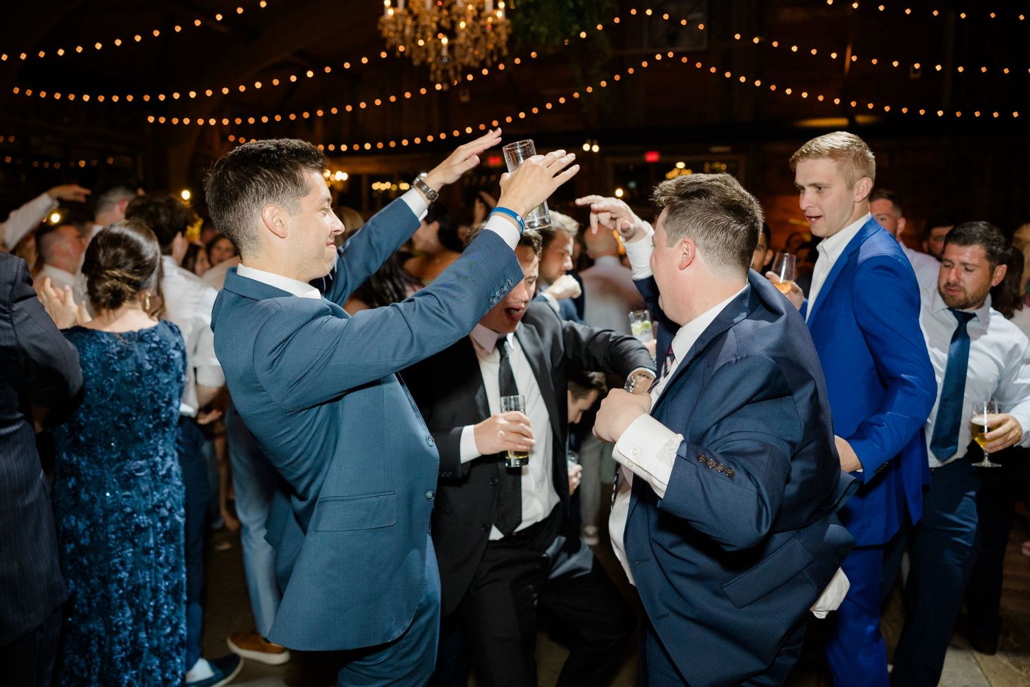 Wedding guests dancing and celebrating on dance floor under hanging string lights and chandelier.