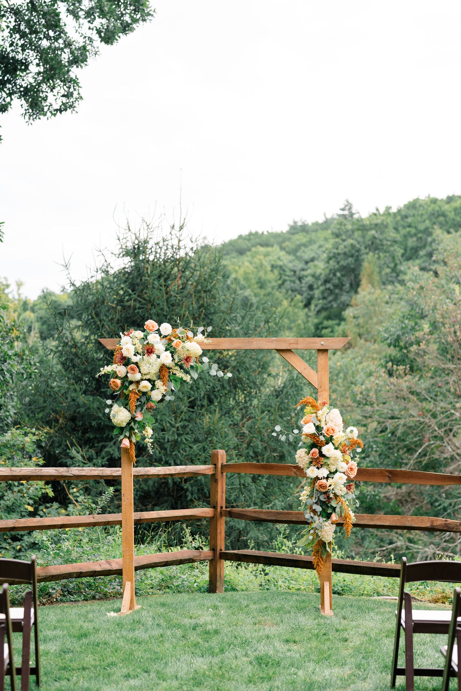 Rustic wooden wedding arch adorned with peach and white floral arrangements set against a lush green forest backdrop.