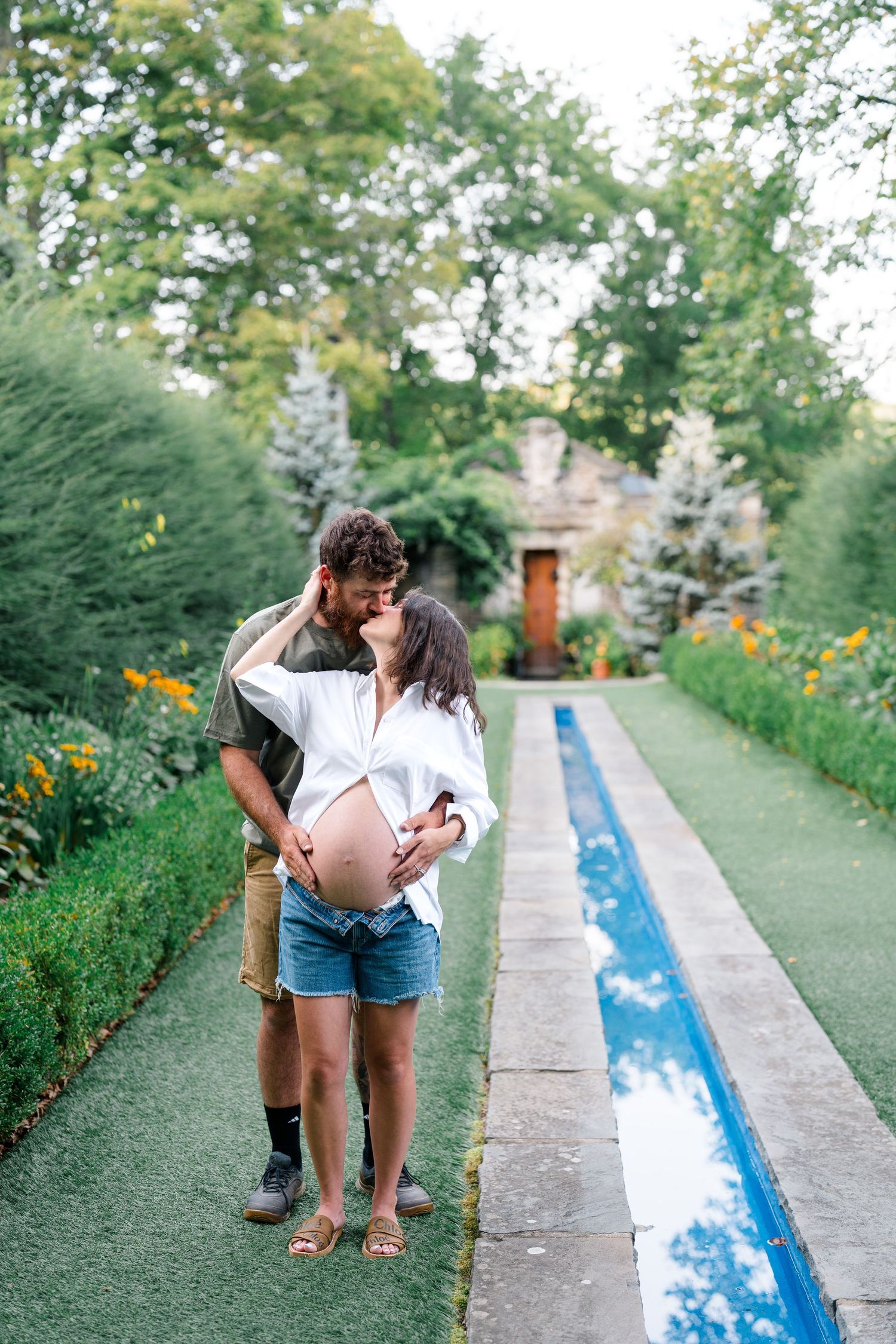 Pregnant couple kissing beside a reflecting pool in lush garden, woman showing baby bump in white shirt.