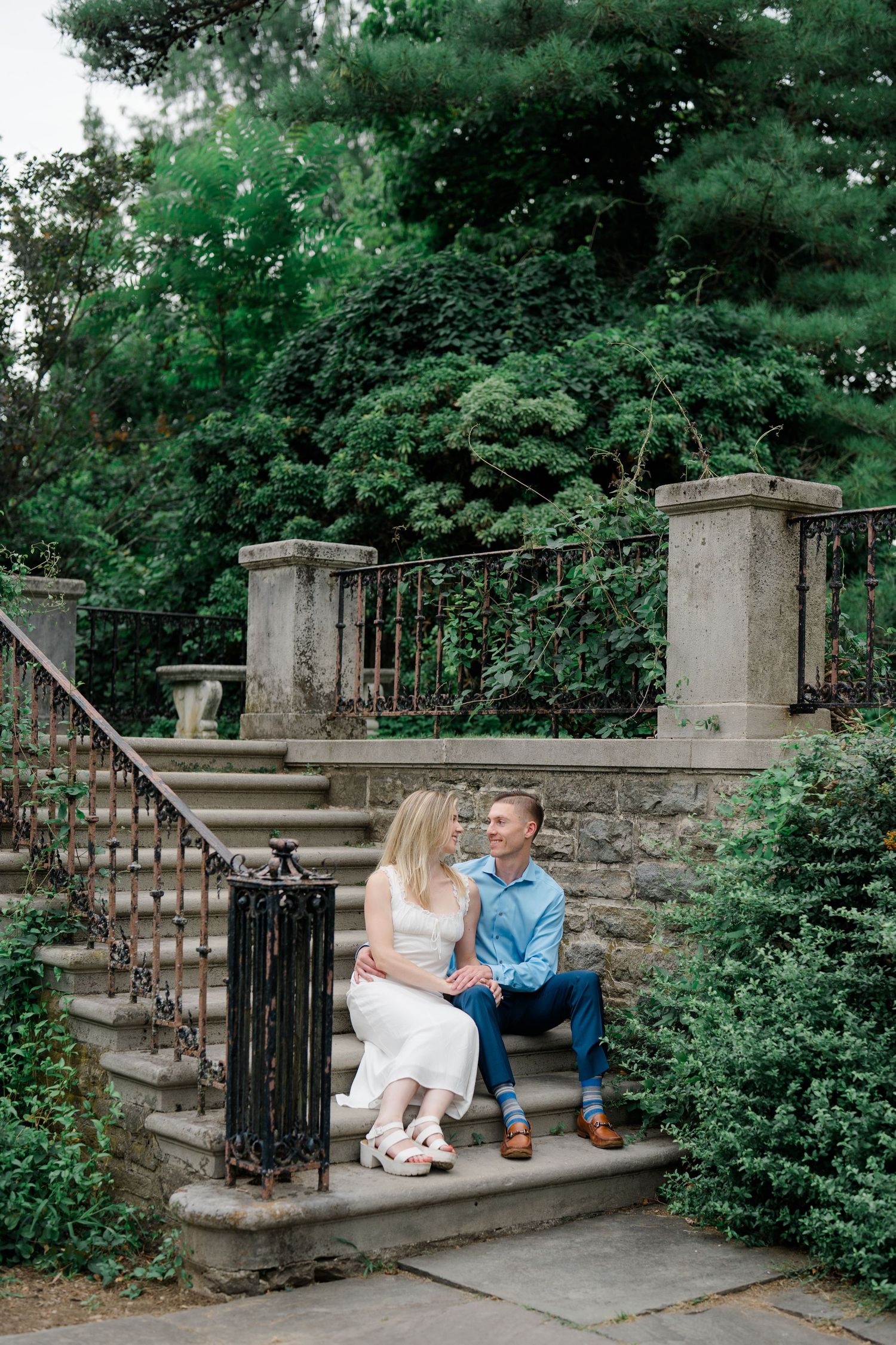 Couple sitting on stone steps surrounded by lush greenery, sharing a romantic moment in a garden setting.