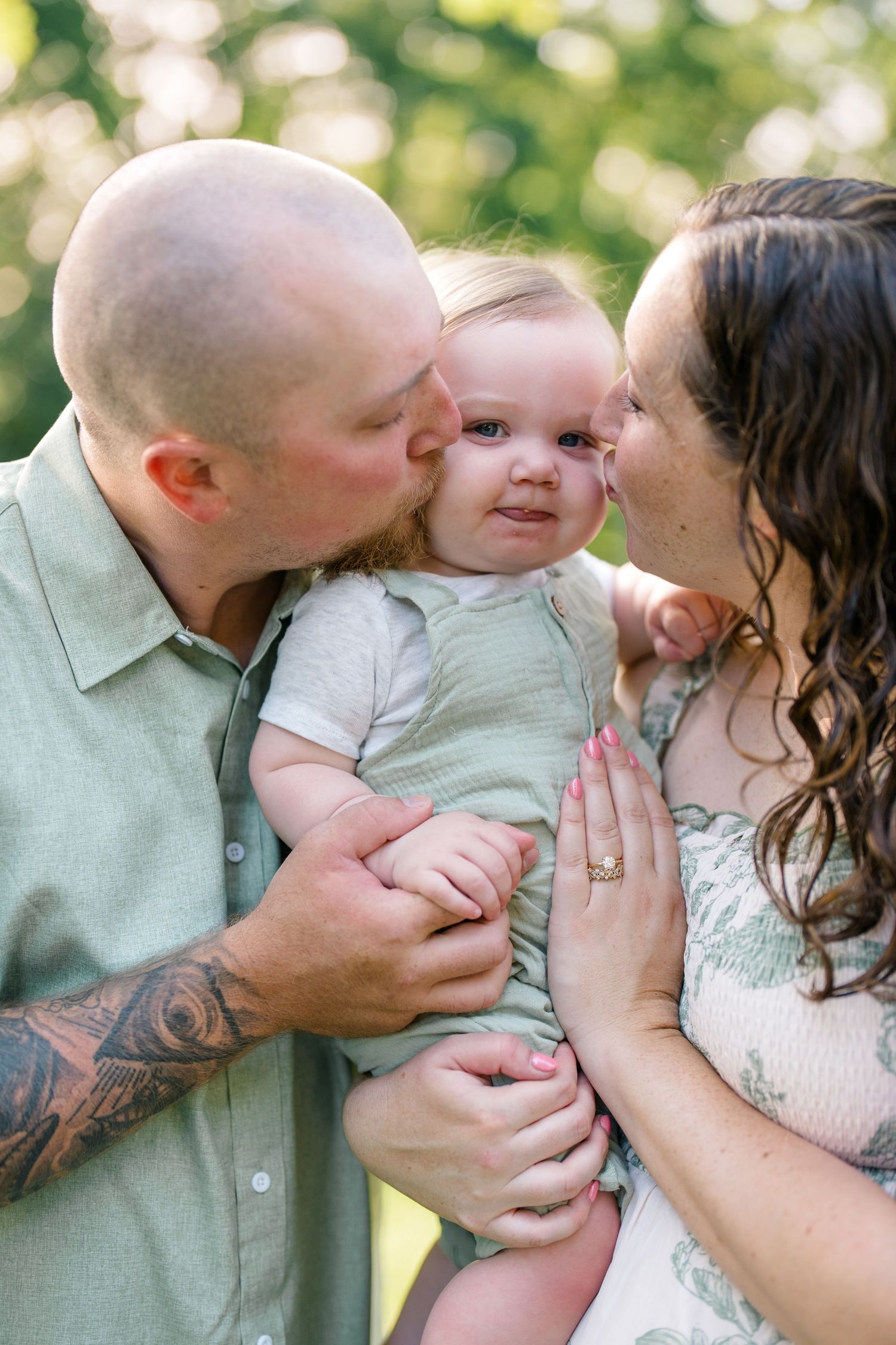 Parents kissing their smiling baby on the cheeks outdoors in a warm, sunlit garden setting.