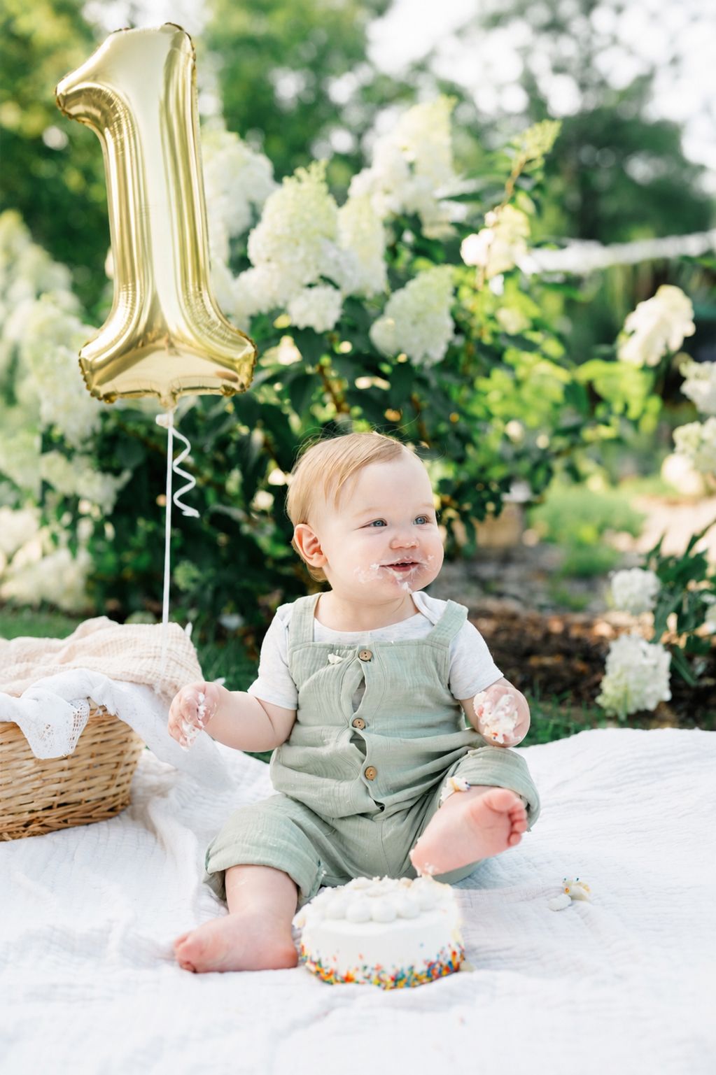 Baby's first birthday cake smash outdoors with gold number one balloon, white flowers, and green romper on white blanket.