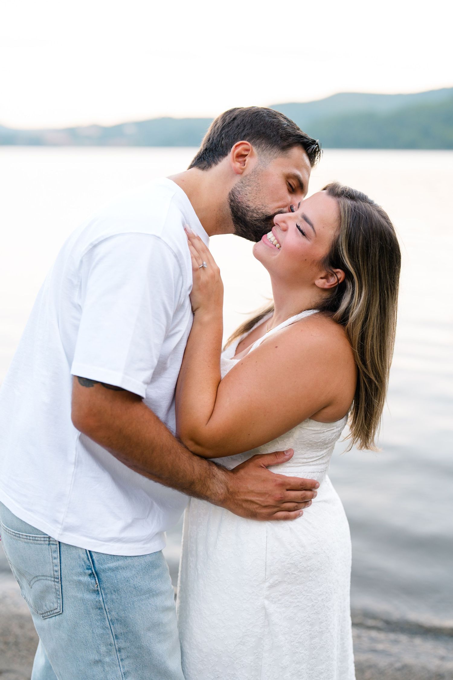 Couple embracing and kissing by a lake, wearing white outfits, sharing a romantic moment on a sandy shore.