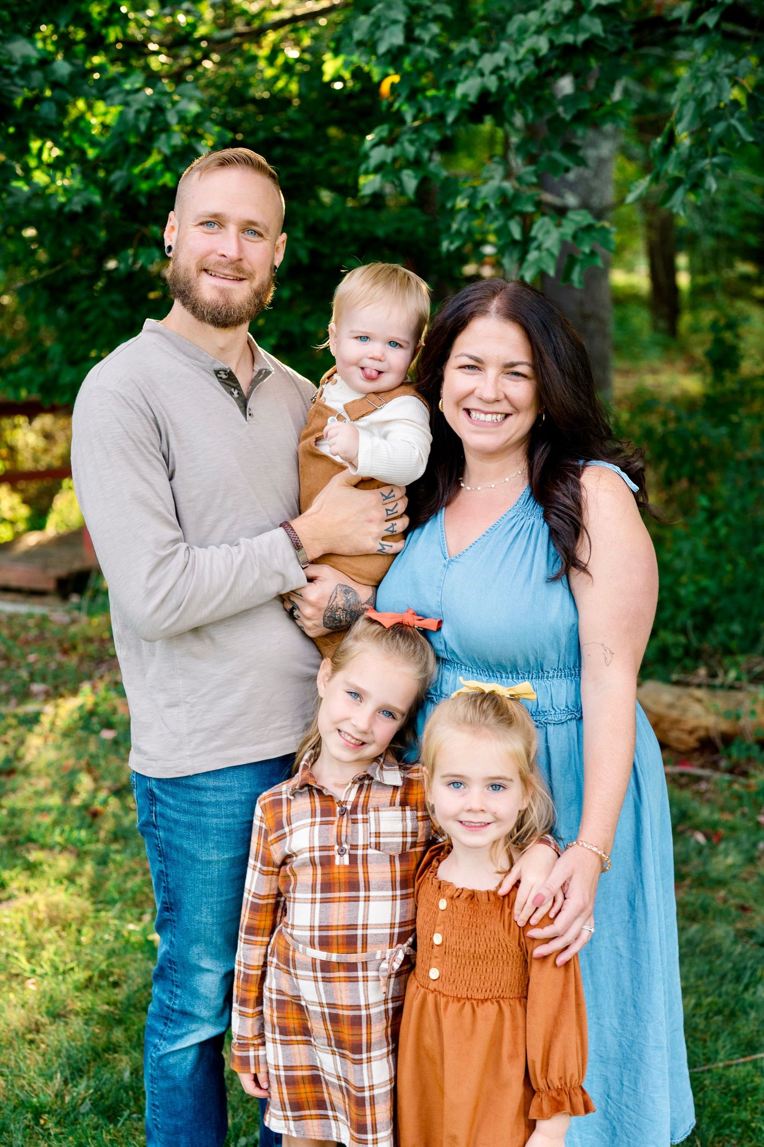 A happy family of five poses outdoors in fall attire, with parents holding three young children amid lush green trees.