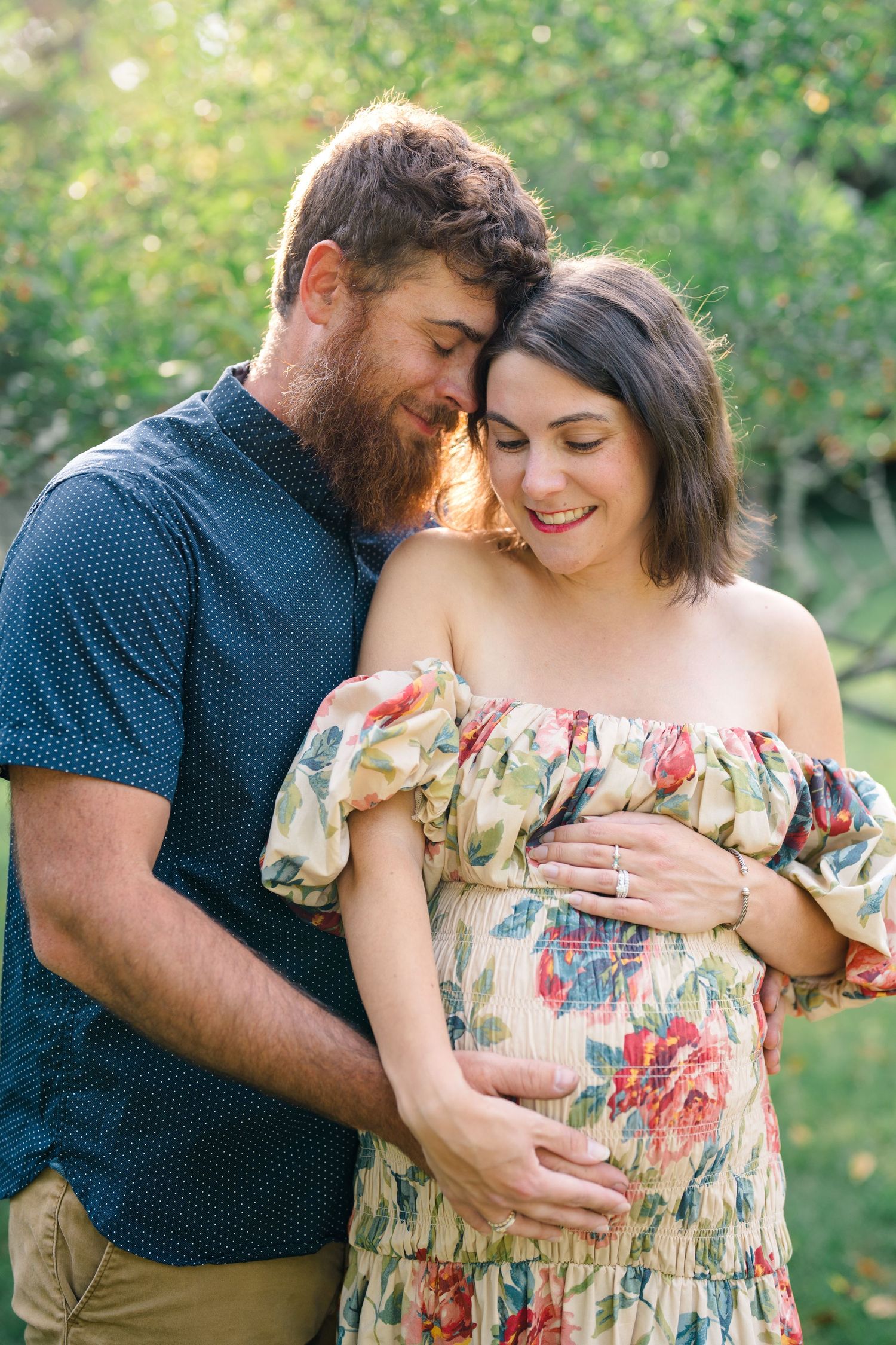 Couple embracing outdoors, pregnant woman in floral off-shoulder dress, man with red beard nuzzling her head in garden.