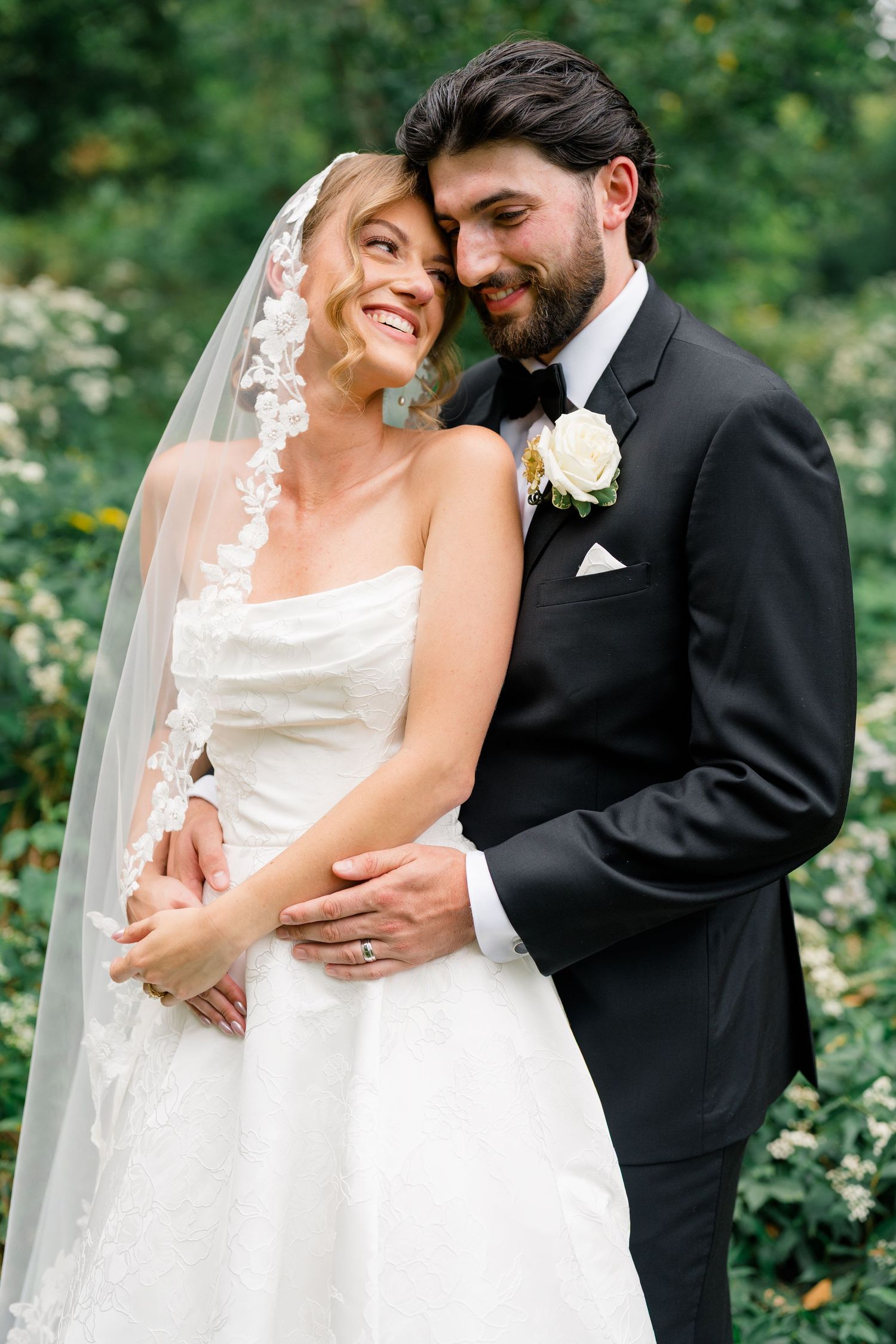 A smiling bride in a white strapless gown and veil embraces a groom in a black tuxedo outdoors.