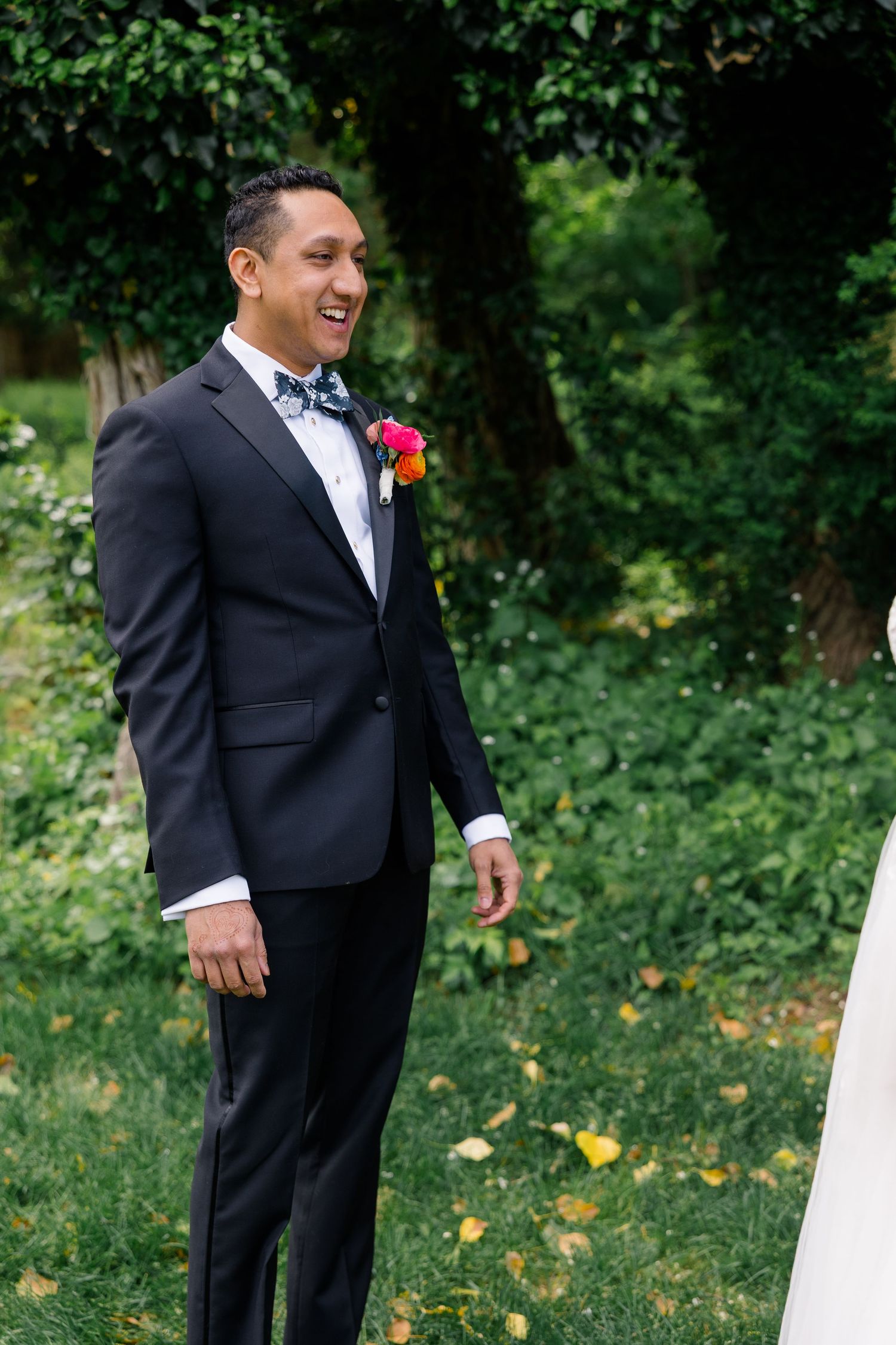 A formal wedding portrait showing a groom in black tuxedo and bowtie with red boutonniere against a garden backdrop.