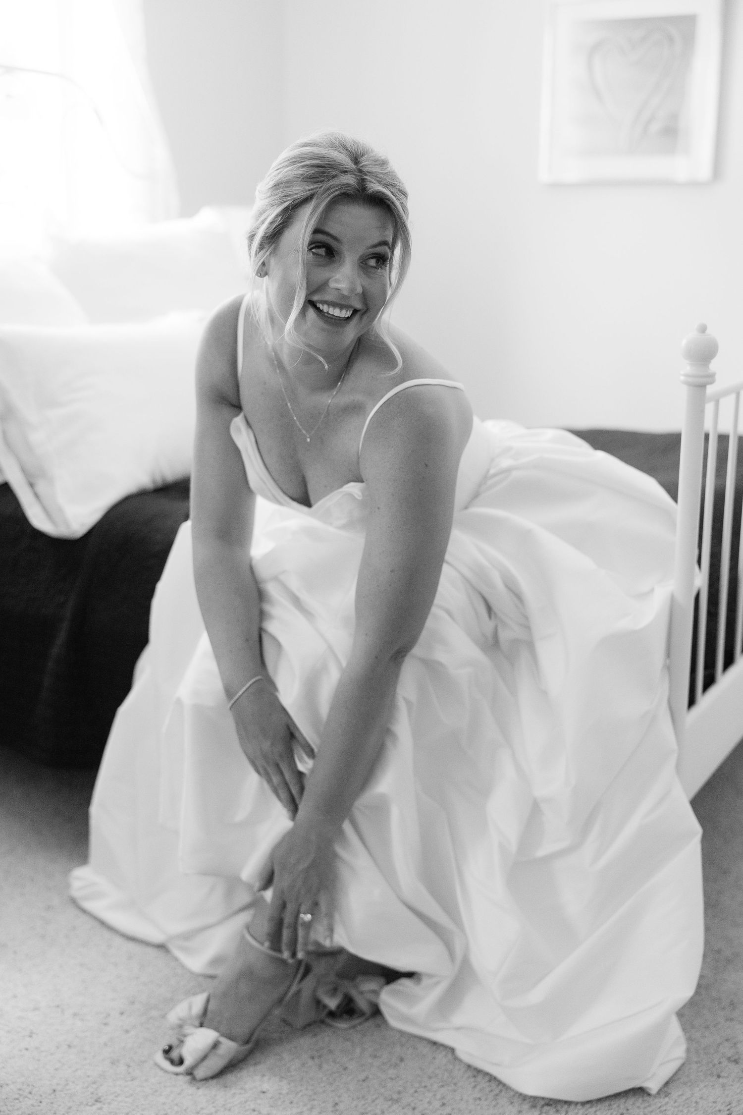 Black and white photo of a smiling bride in a white gown putting on her heels.