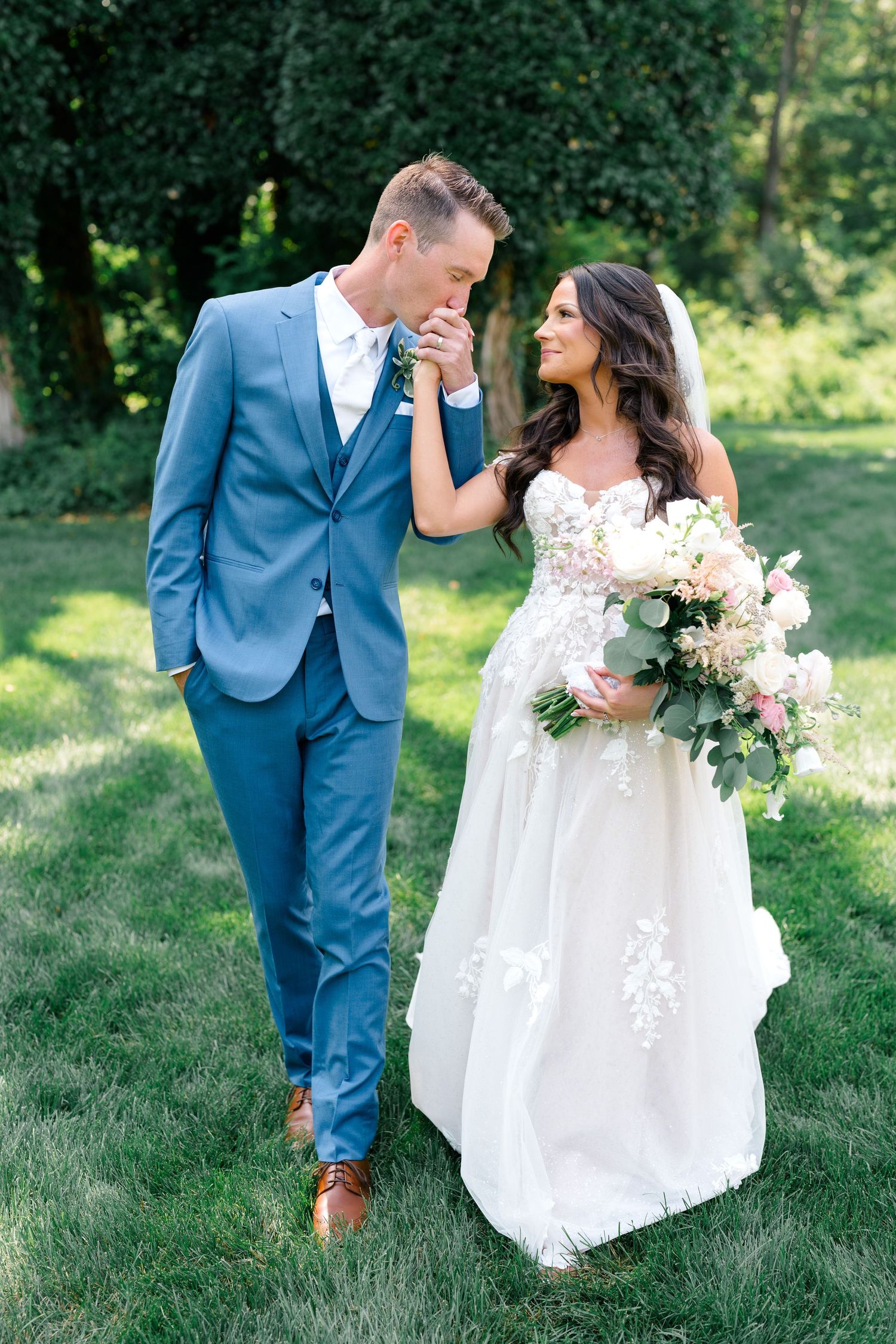 A romantic garden wedding portrait with couple in blue suit and white dress holding pink bouquet.