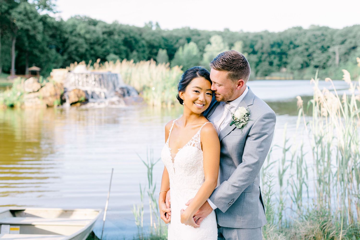 Romantic couple embracing by a scenic lake with tall grass and trees in the background at sunset.