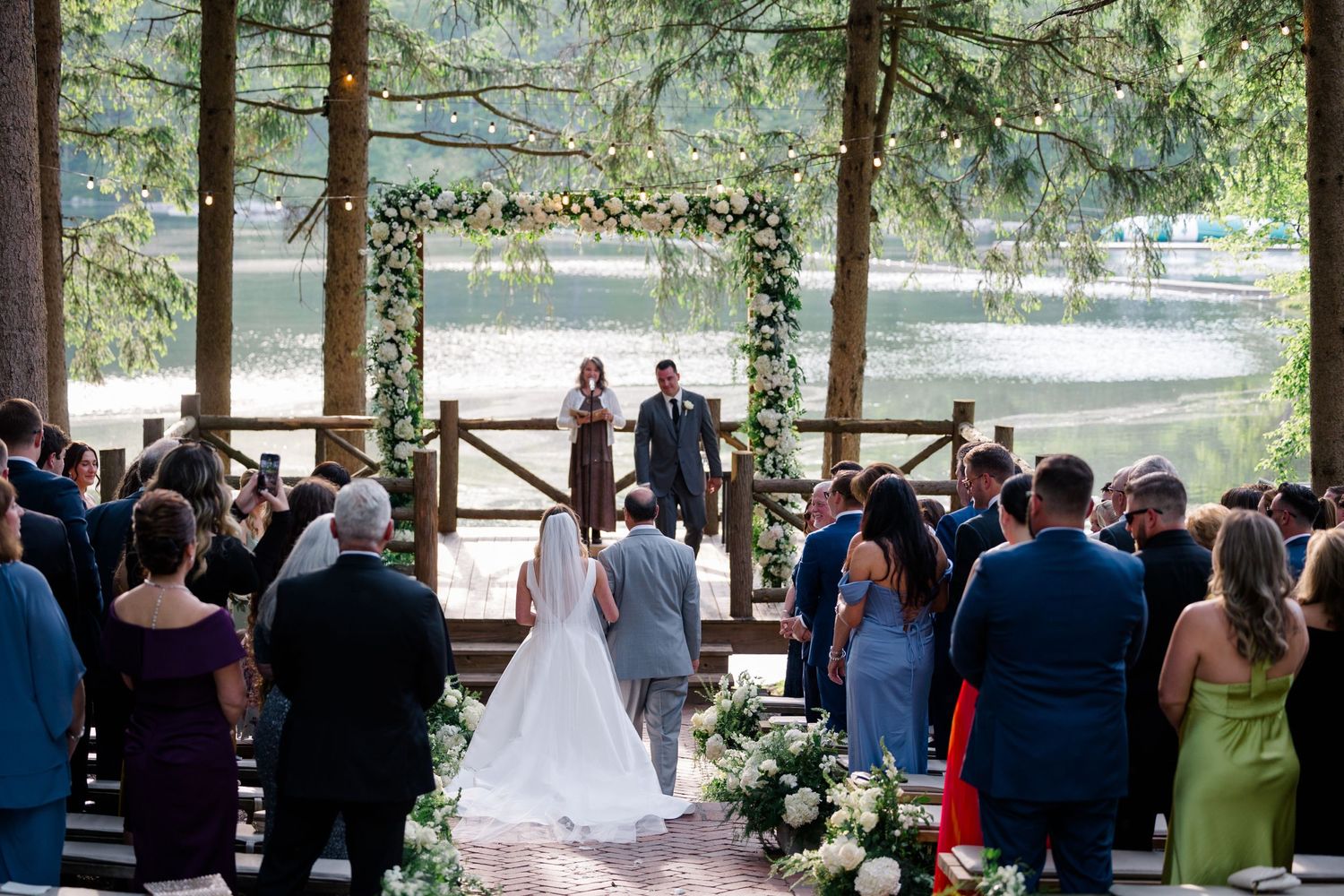 Outdoor wedding ceremony on a rustic wooden deck decorated with floral garlands overlooking a scenic lake in the woods.