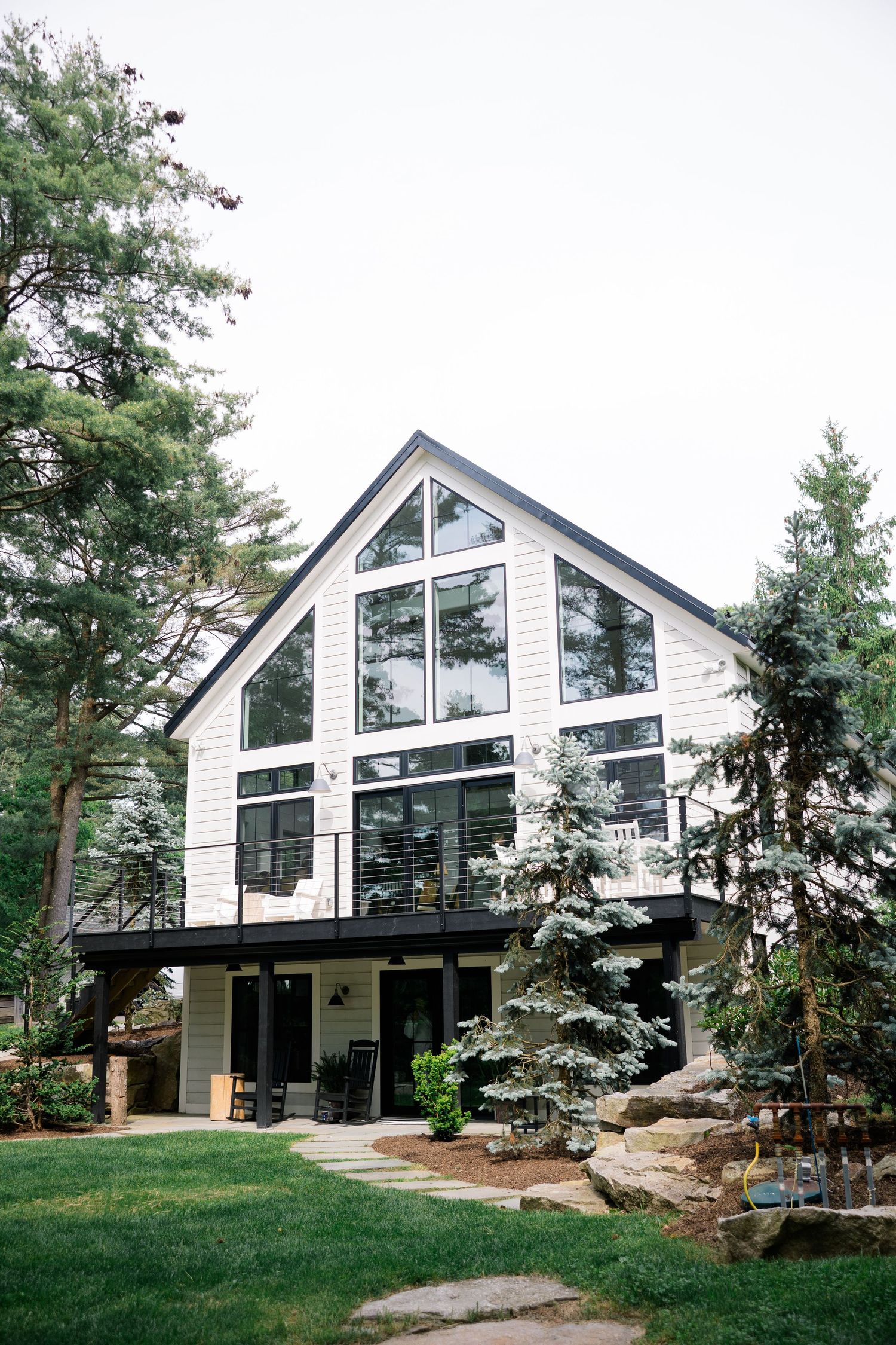 Modern A-frame house with large windows and glass facade nestled among evergreen trees with landscaped rock garden.
