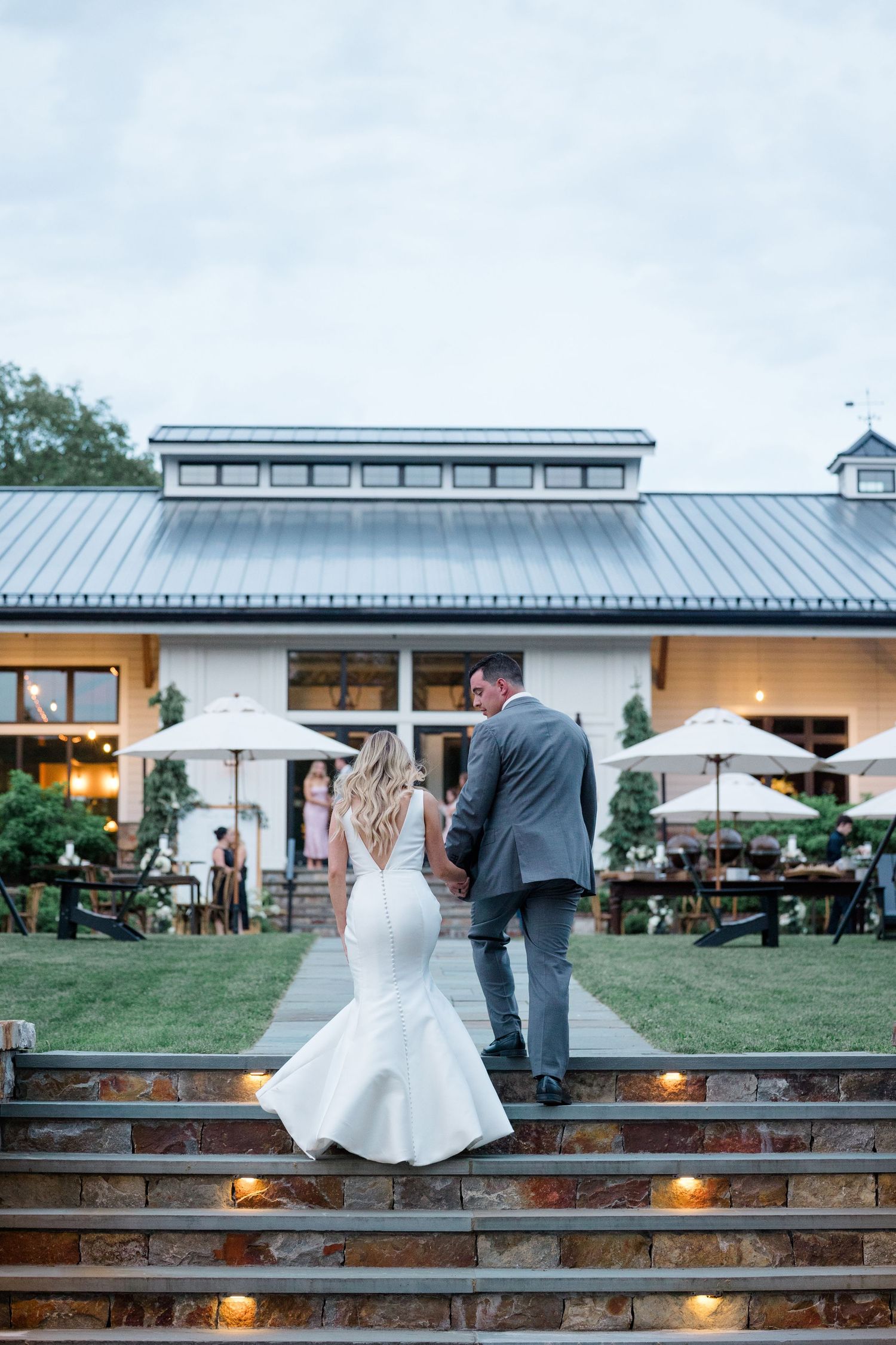 Couple stands on stone steps outside a white building with a metal roof and landscaped garden.
