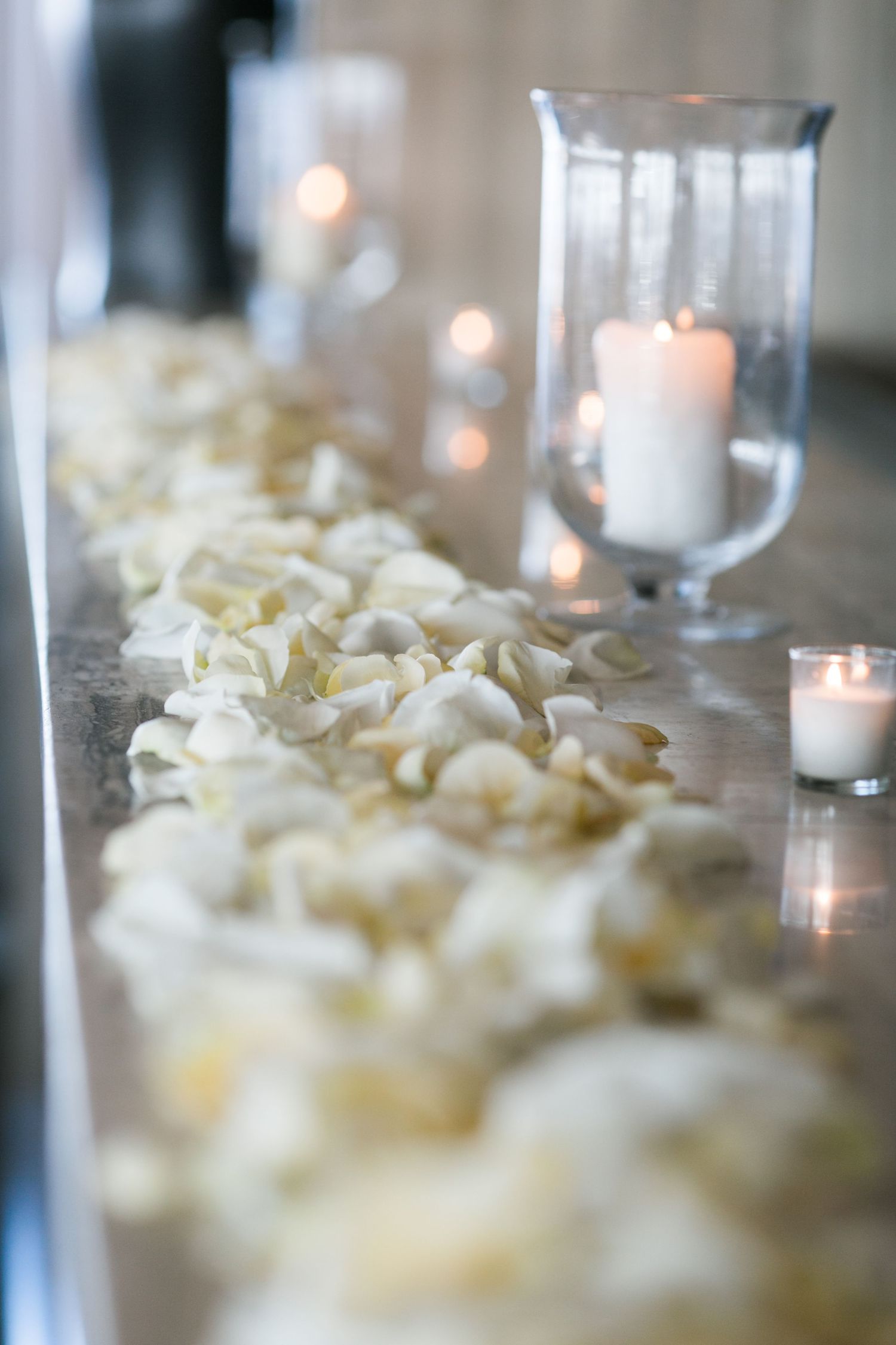 White rose petals scattered along a table with glowing candles in glass hurricane holders create a romantic atmosphere.