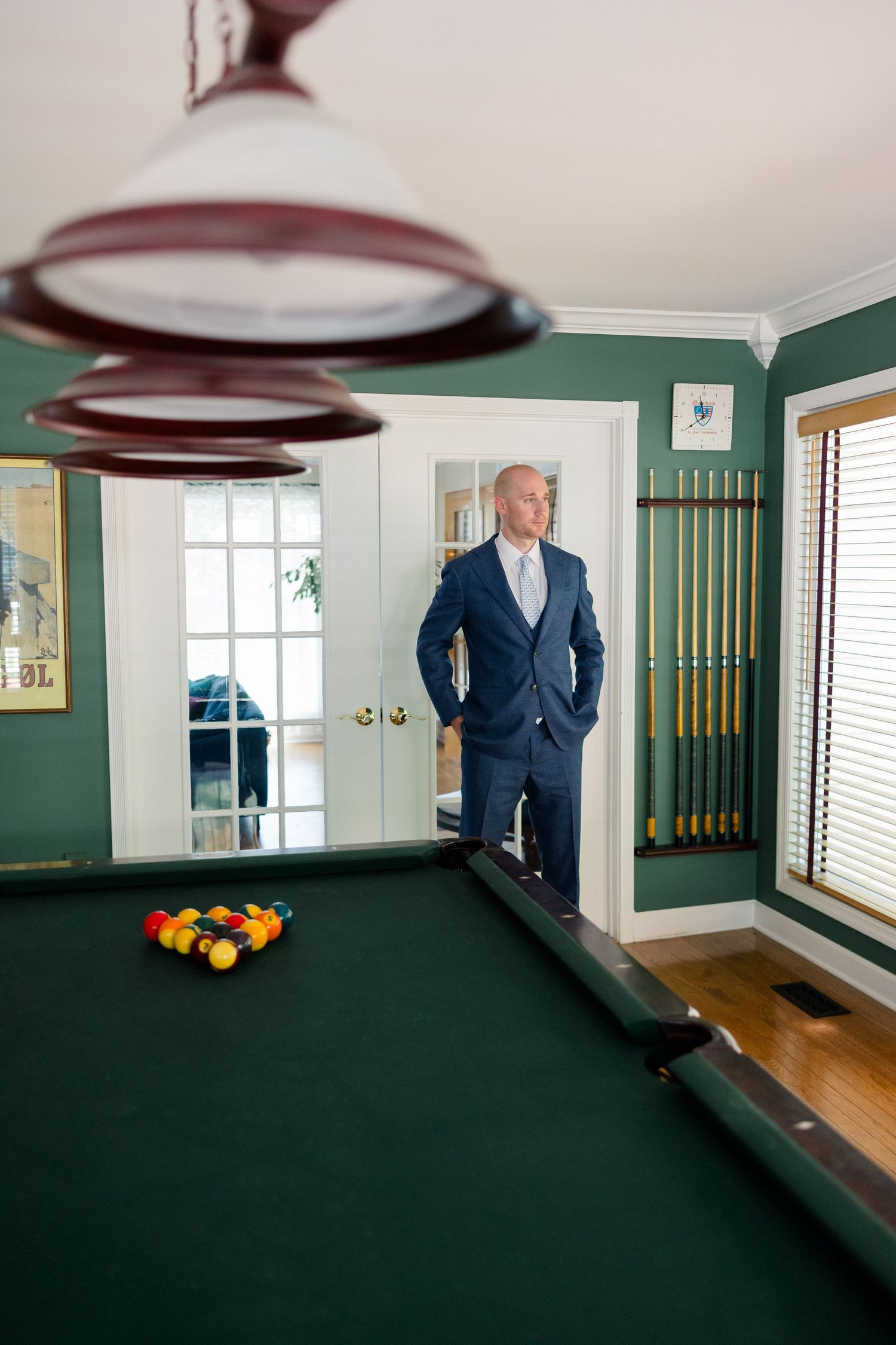 Man in navy suit standing by a billiard table in a green game room with pool cues mounted on the wall.