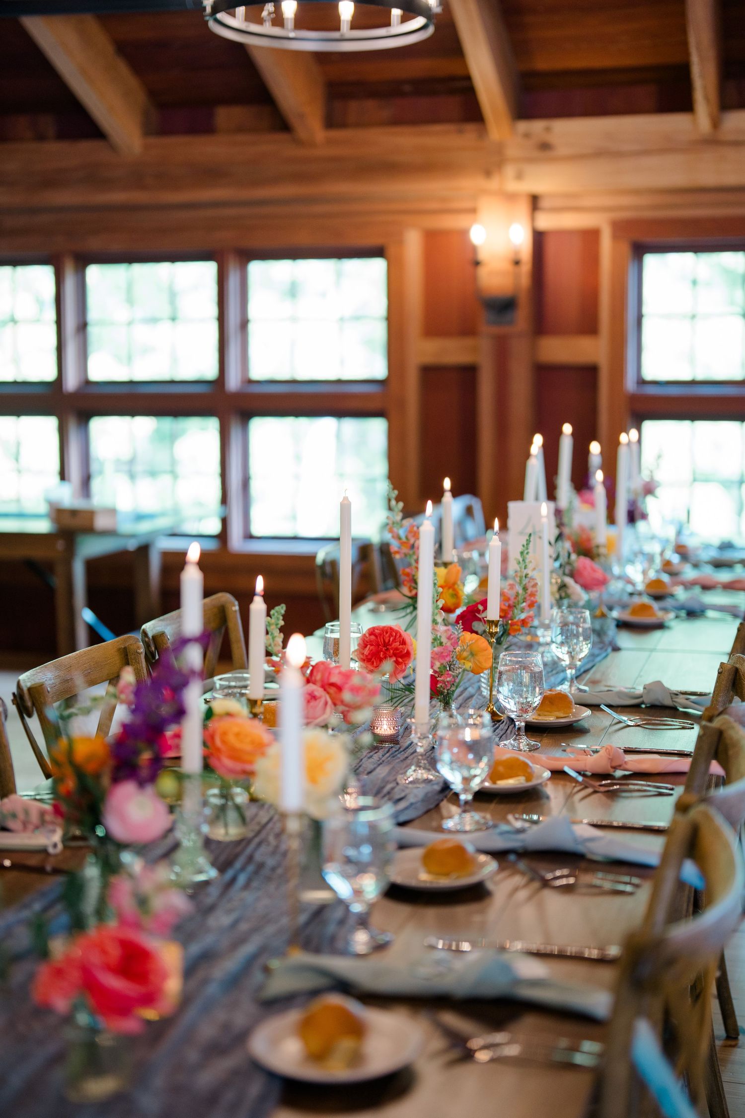 Elegant wedding reception table setting with colorful floral centerpieces and tall white candles in rustic venue.