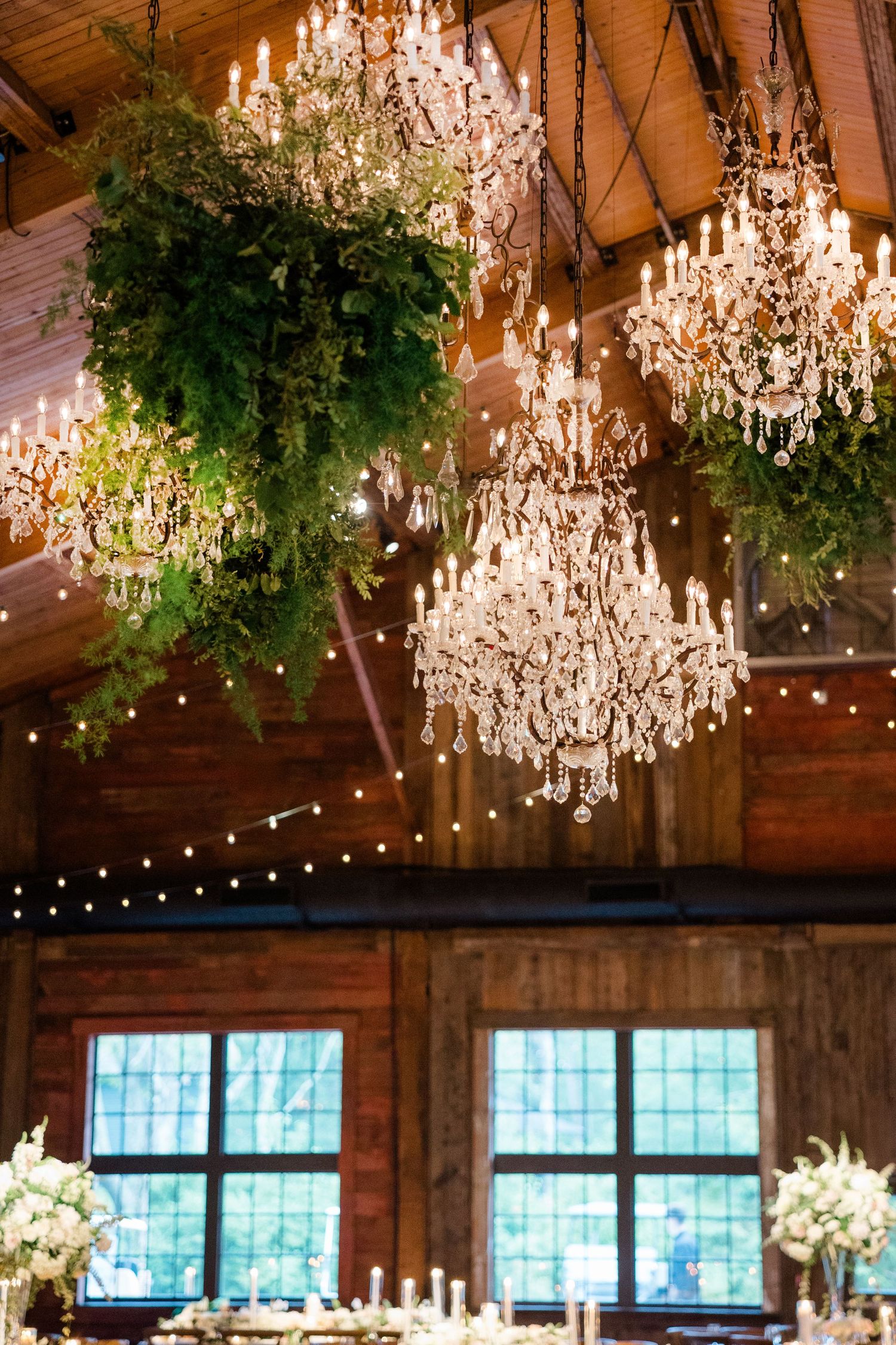 Crystal chandeliers and hanging greenery create an elegant rustic barn wedding reception ceiling.