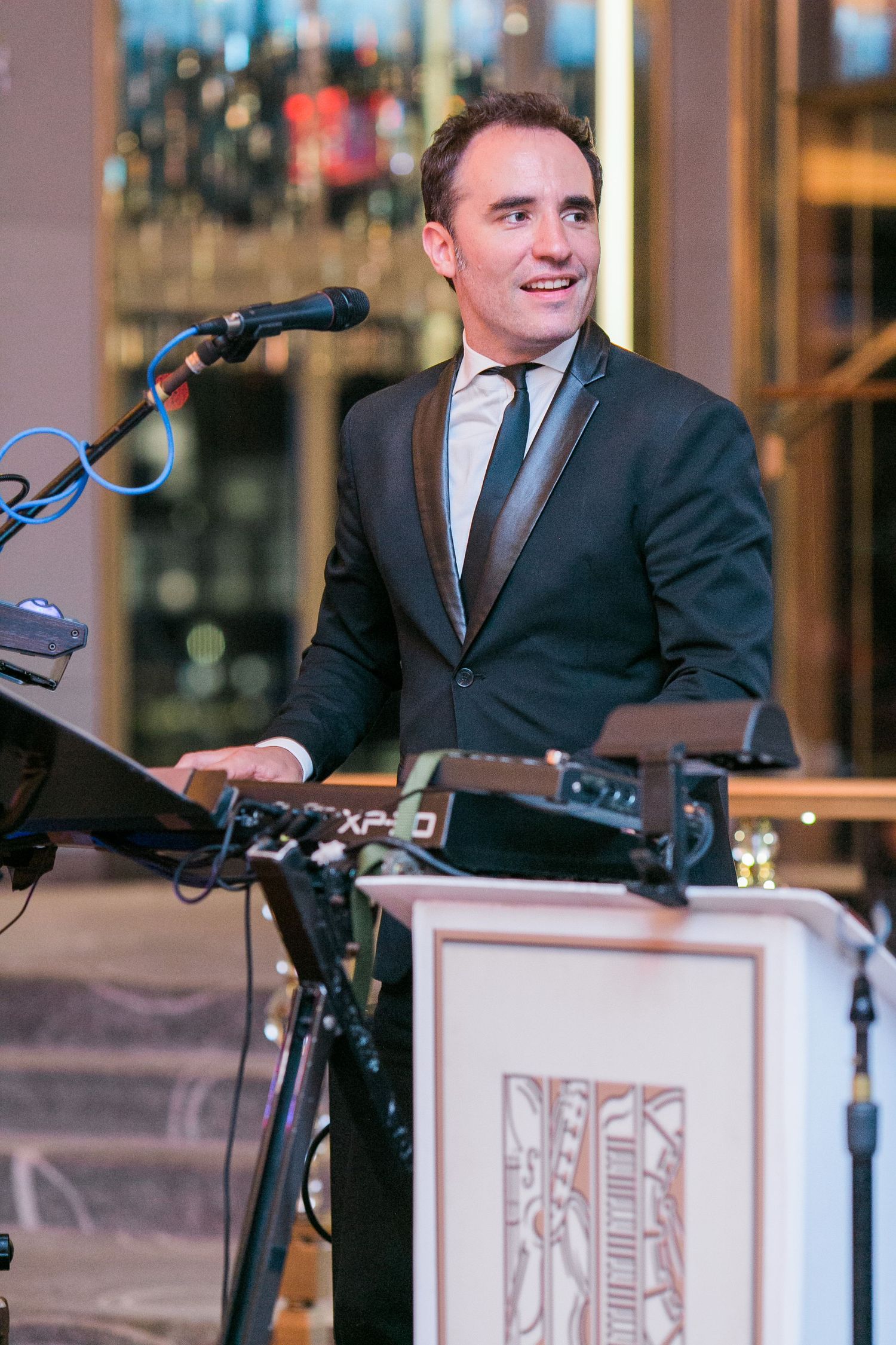 A performer in a dark suit and tie stands smiling behind event equipment with a microphone setup at a venue.