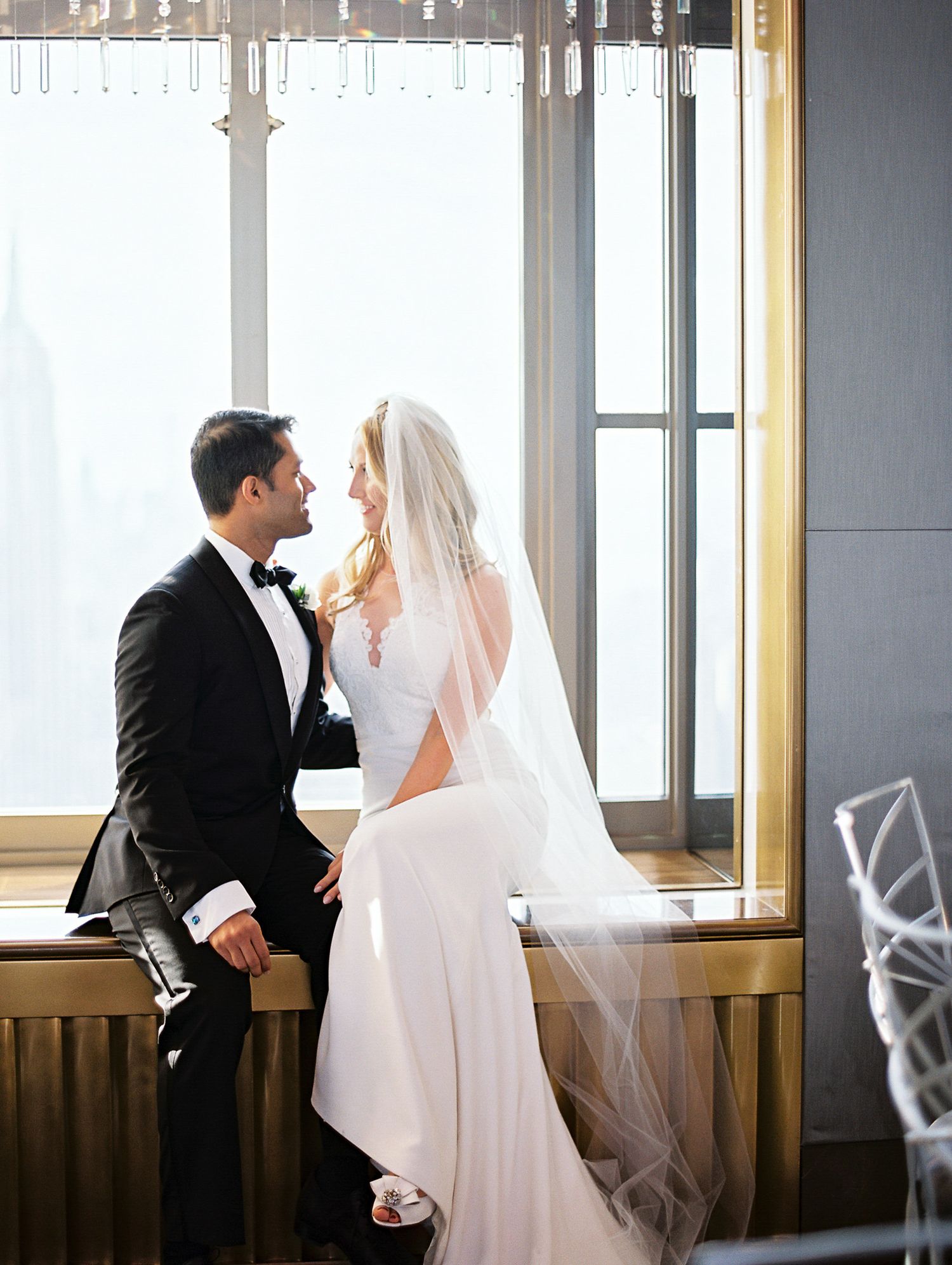 A couple in wedding attire shares an intimate moment by a large window in an elegant indoor setting.