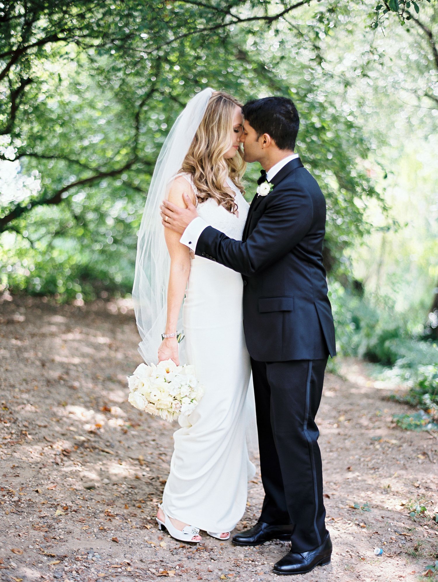Bride and groom share an intimate kiss beneath green trees while holding a white bouquet.