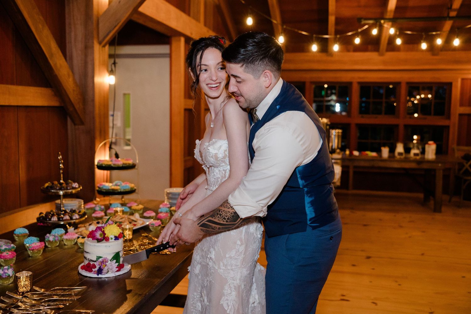 A couple cuts their wedding cake together in a warmly lit rustic wooden reception venue.