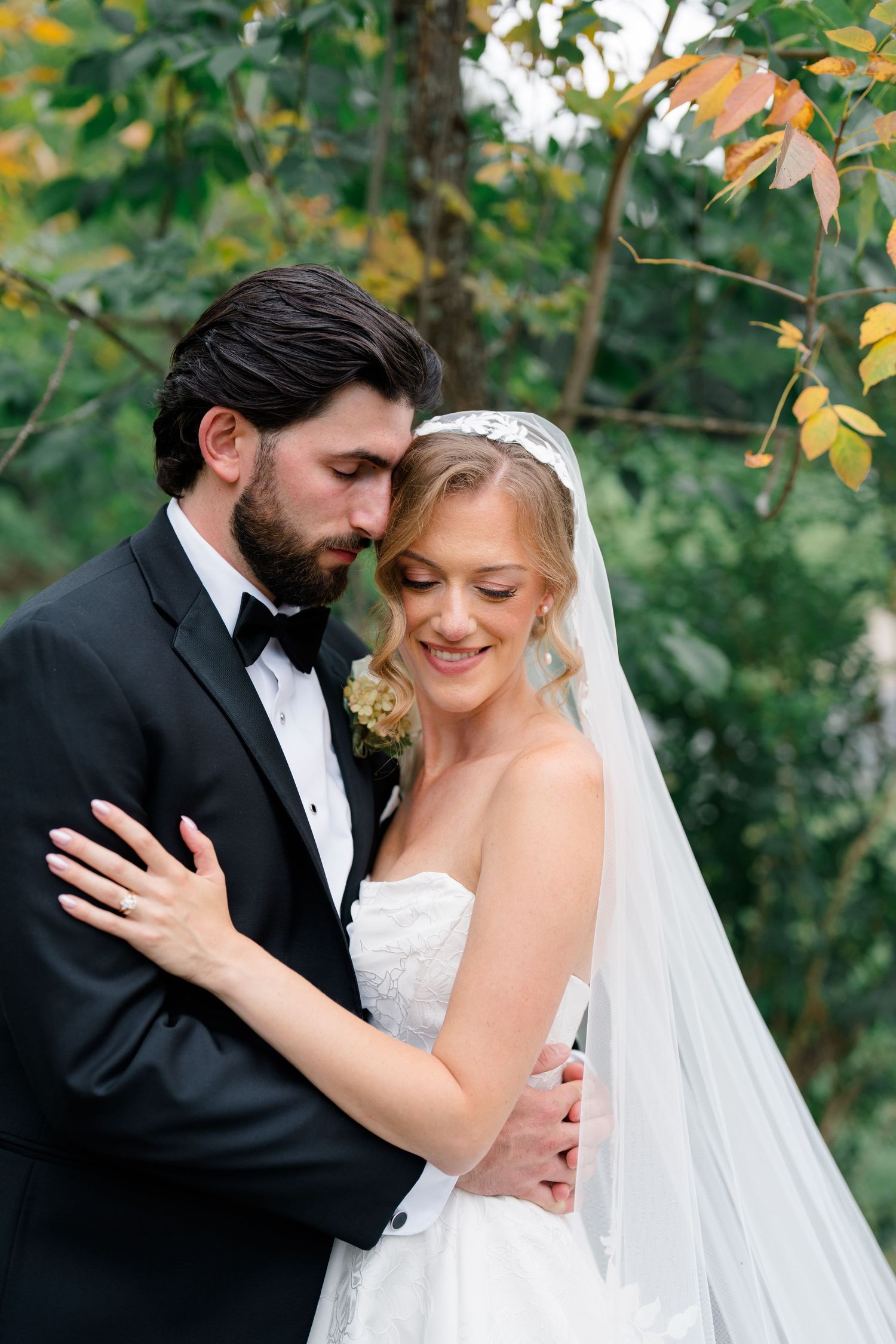 Bride and groom embrace outdoors in autumn, groom in tuxedo, bride in strapless gown with veil, surrounded by fall foliage.
