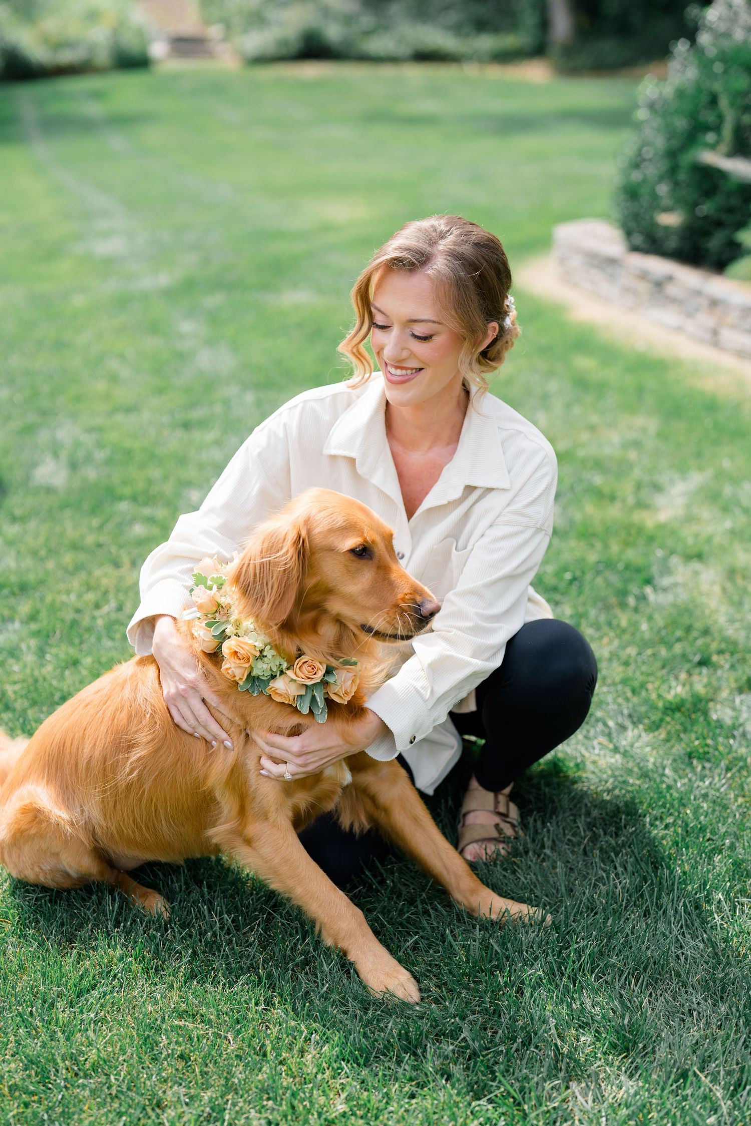 Smiling bride in white shirt poses with a golden retriever wearing a floral flower crown on green grass.