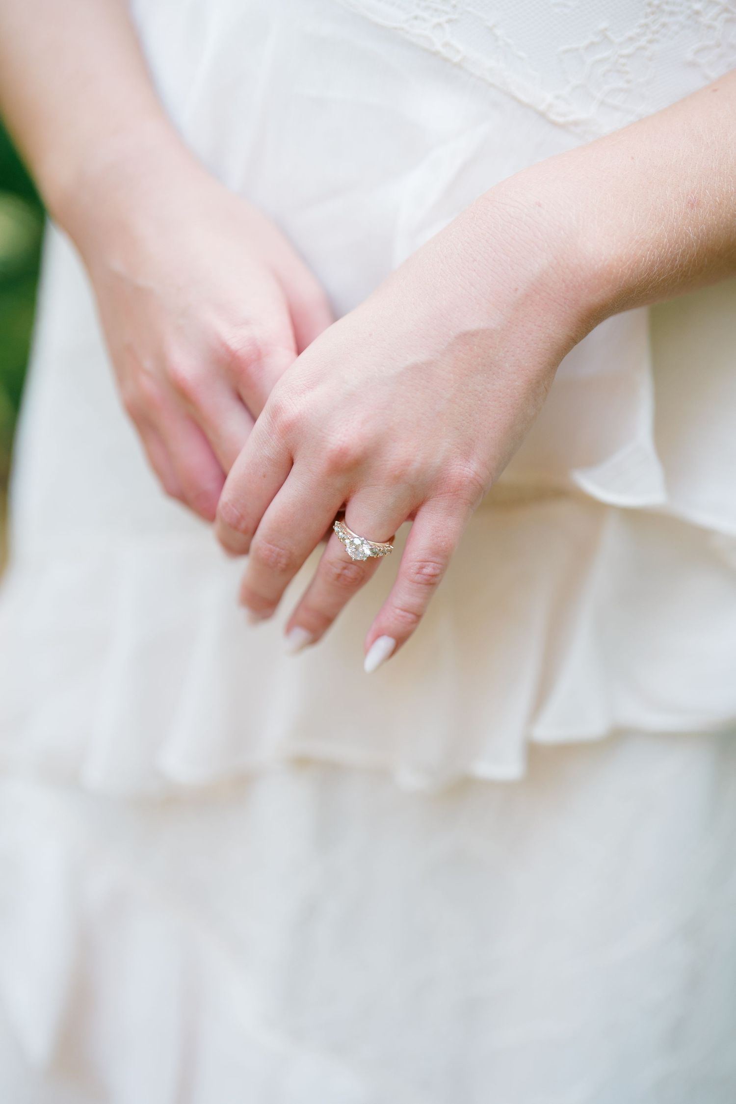 Close-up of a bride's hands showing a delicate diamond engagement ring on her finger against a white dress.