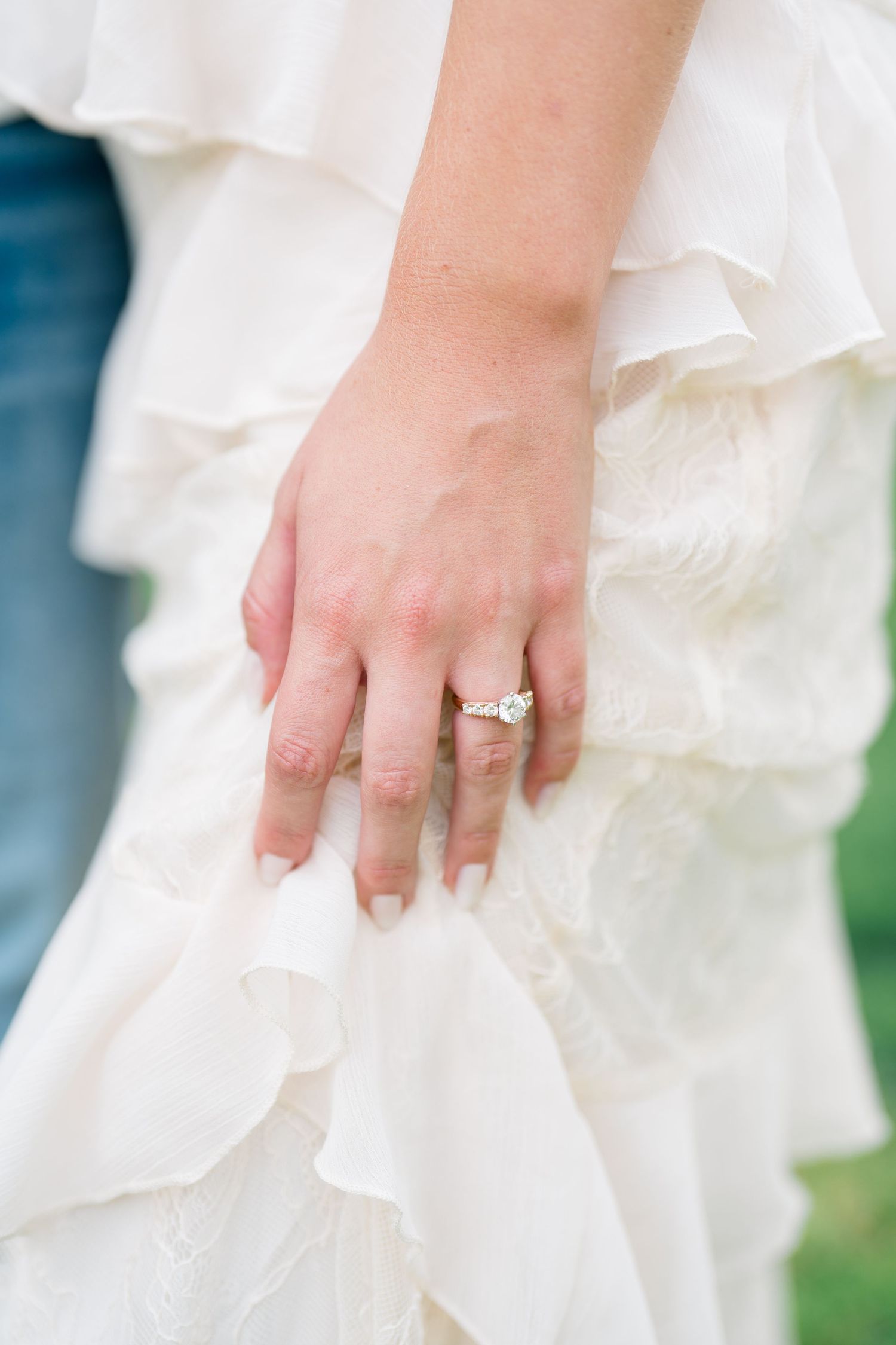 Close-up of a bride's hand resting on her white ruffled wedding dress, showcasing a sparkling diamond engagement ring.
