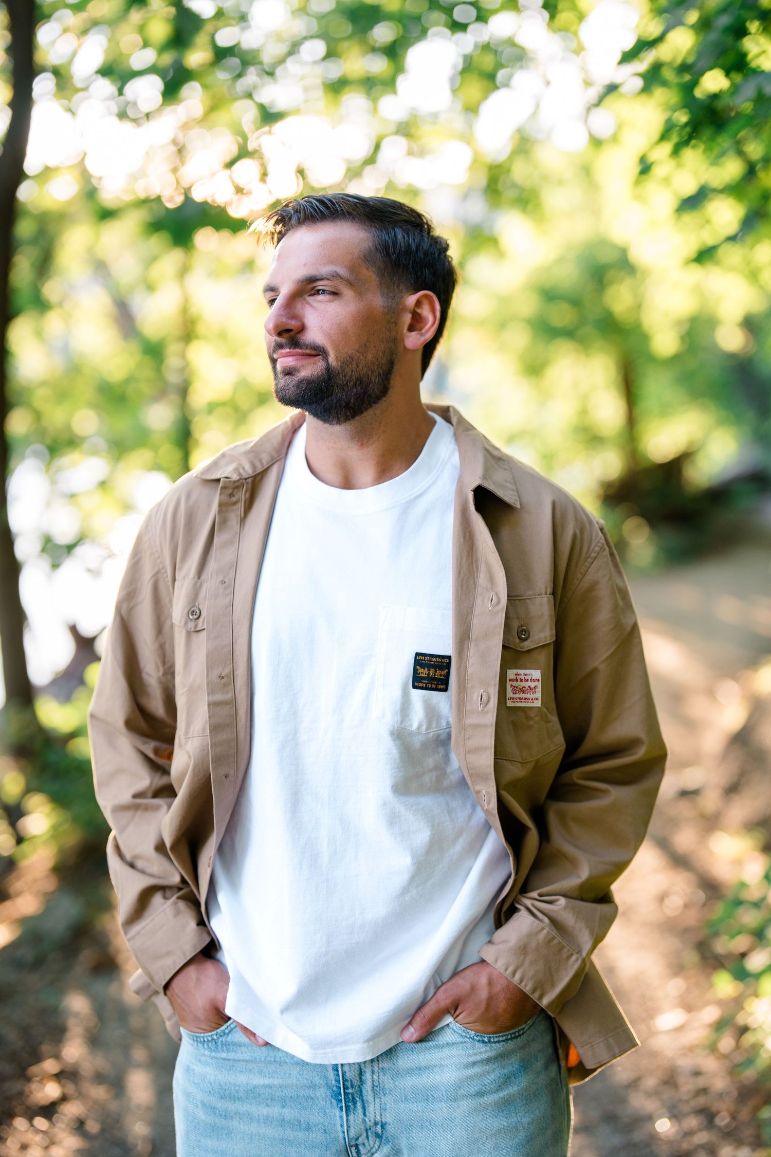 Bearded man in white tee and tan jacket stands outdoors among green trees, hands in pockets.