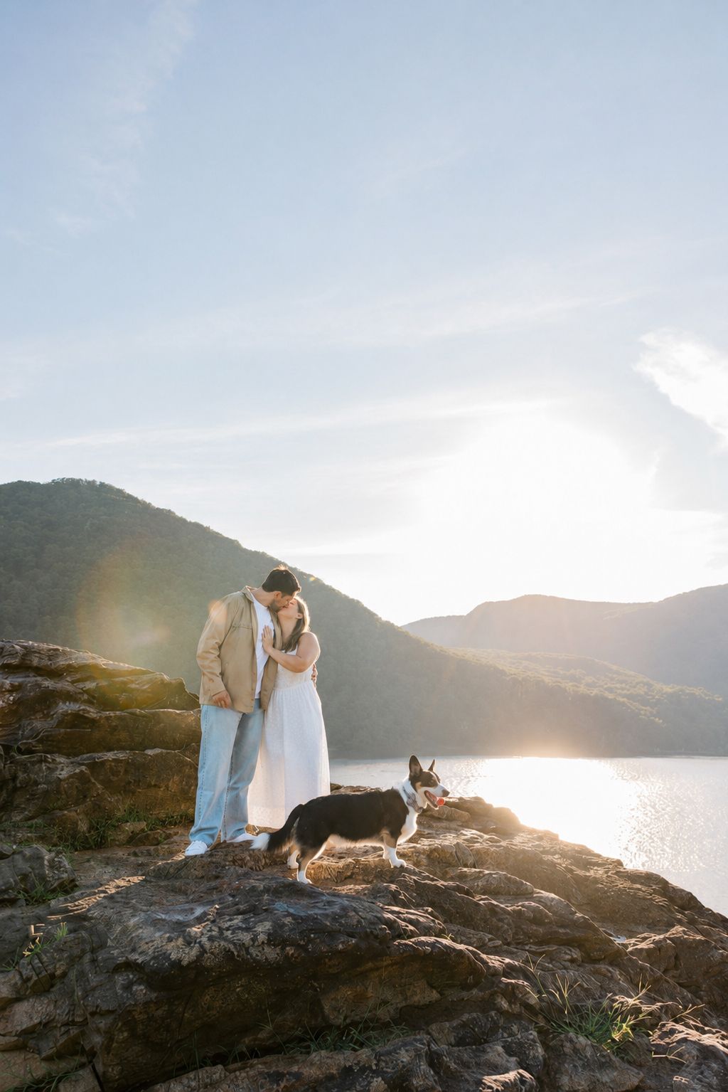 Couple in wedding attire sharing a romantic moment on rocky overlook with dog, mountain lake and golden sunset backdrop.