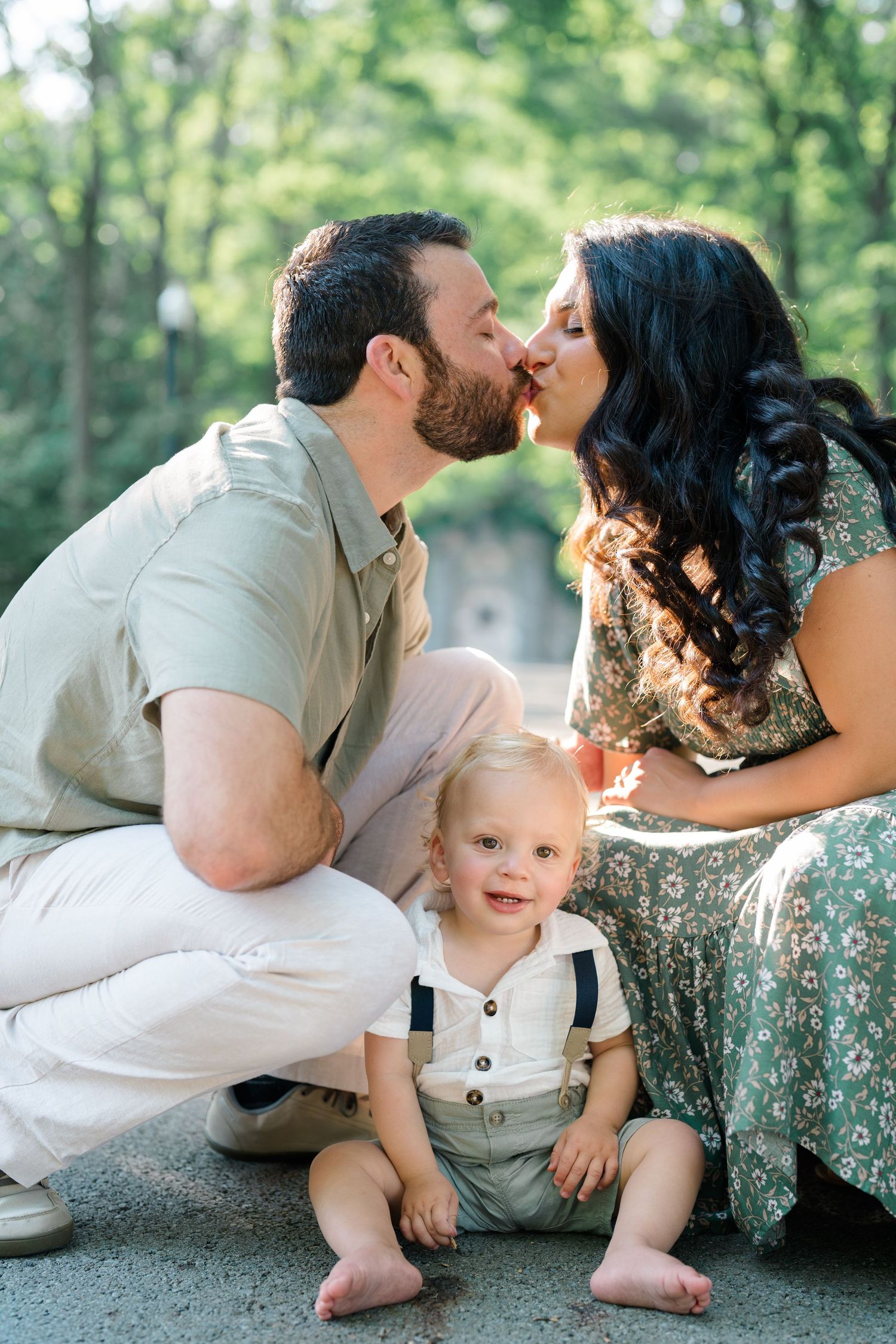 A couple shares a kiss while their smiling toddler sits in front of them outdoors in a lush green setting.