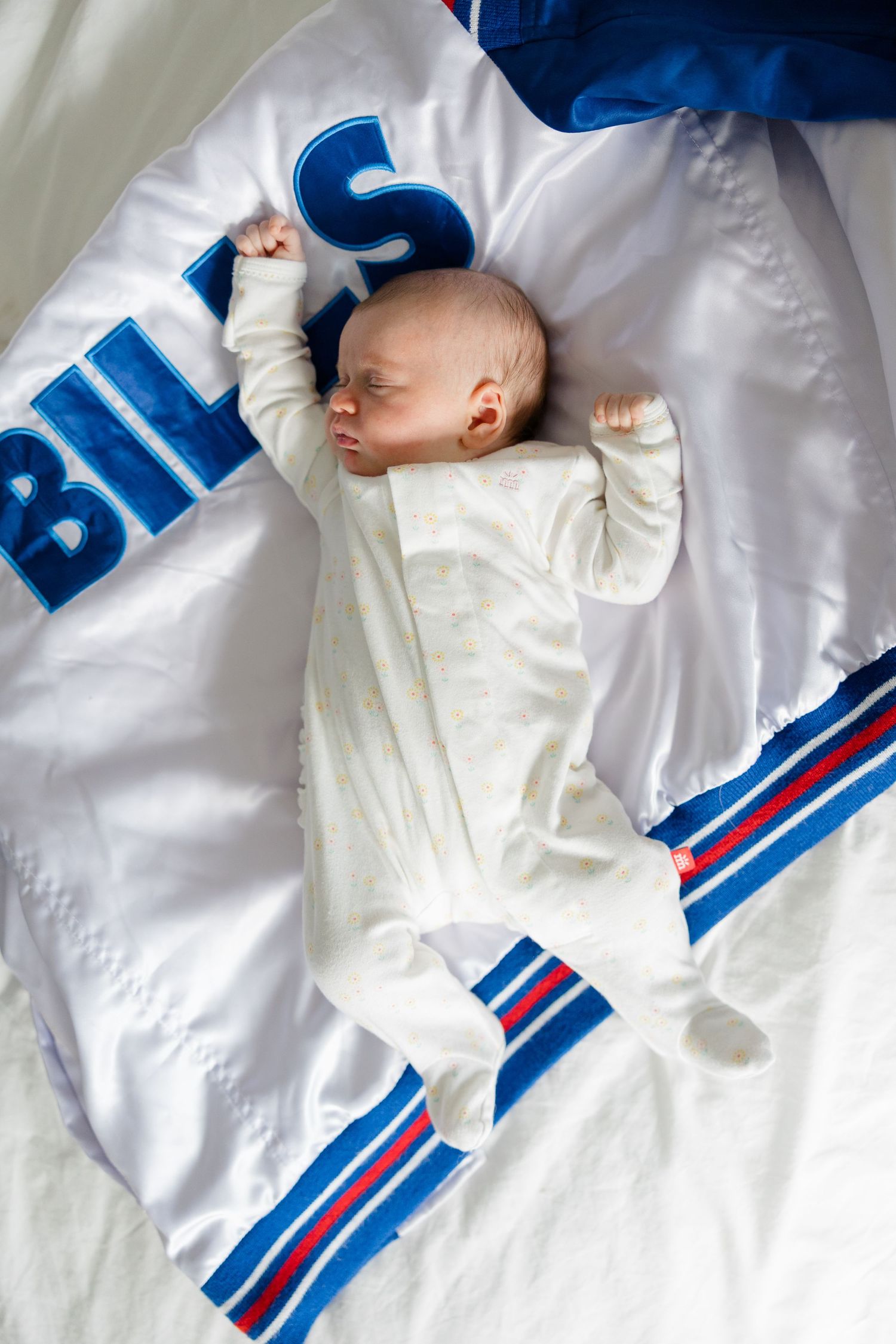 Baby in white sleeper lying on a Buffalo Bills football team blanket with blue and red stripes.