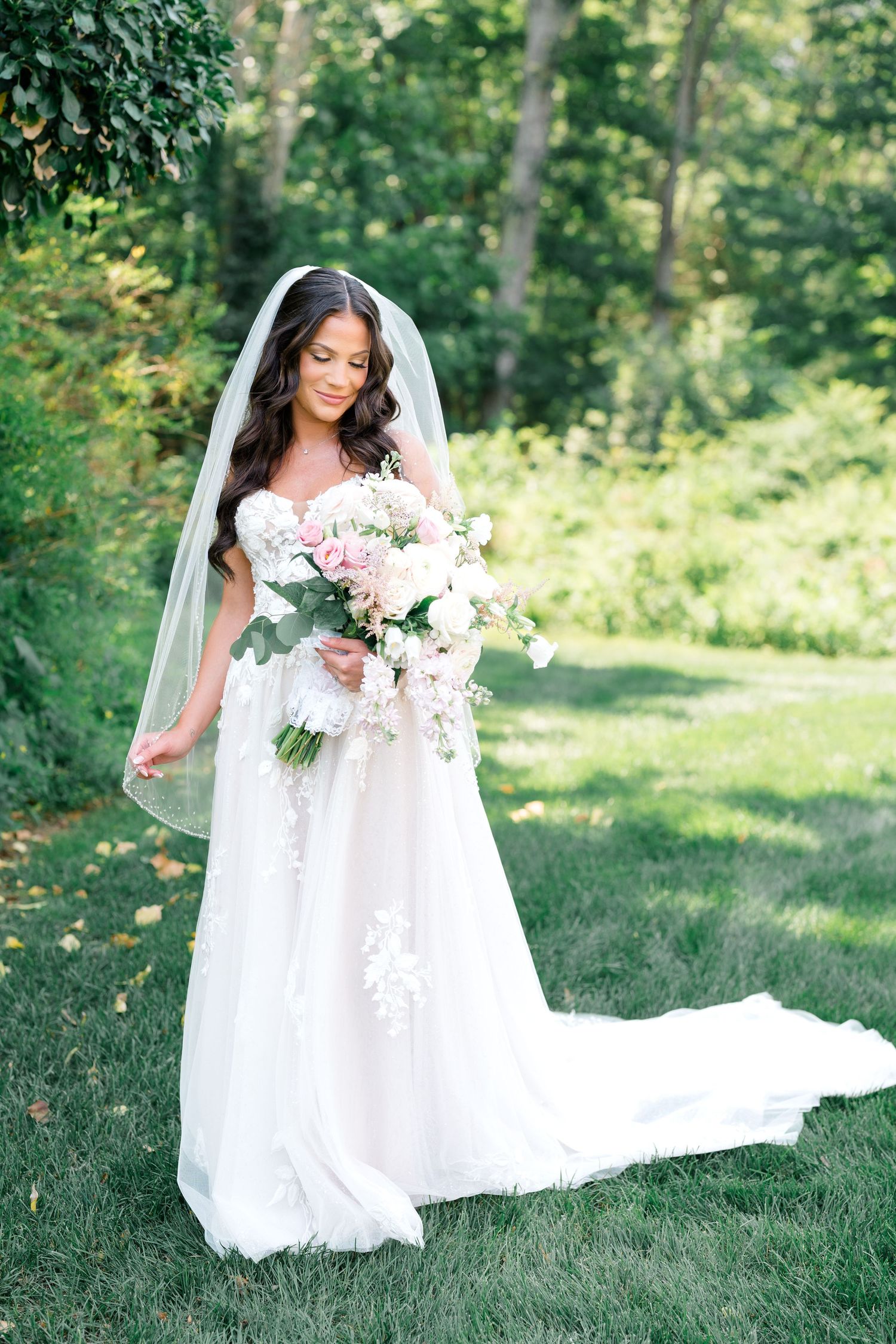 A bride in a flowing white wedding gown poses outdoors in a garden setting while holding a pink and white floral bouquet.