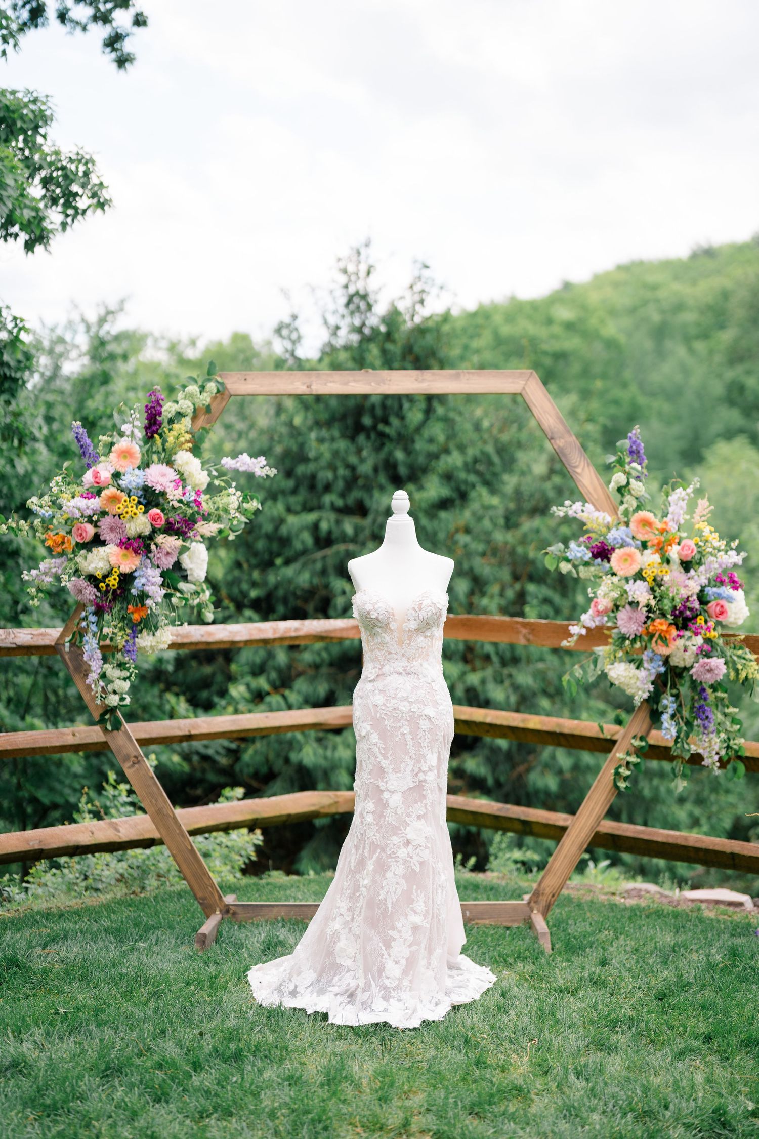 White lace wedding dress displayed outdoors with hexagonal arch and colorful floral arrangements on grassy hill.