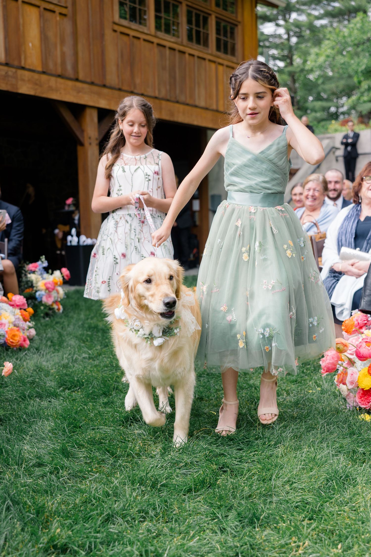 Young attendants in vintage-style dresses walk with golden retriever down flower-lined outdoor wedding aisle.