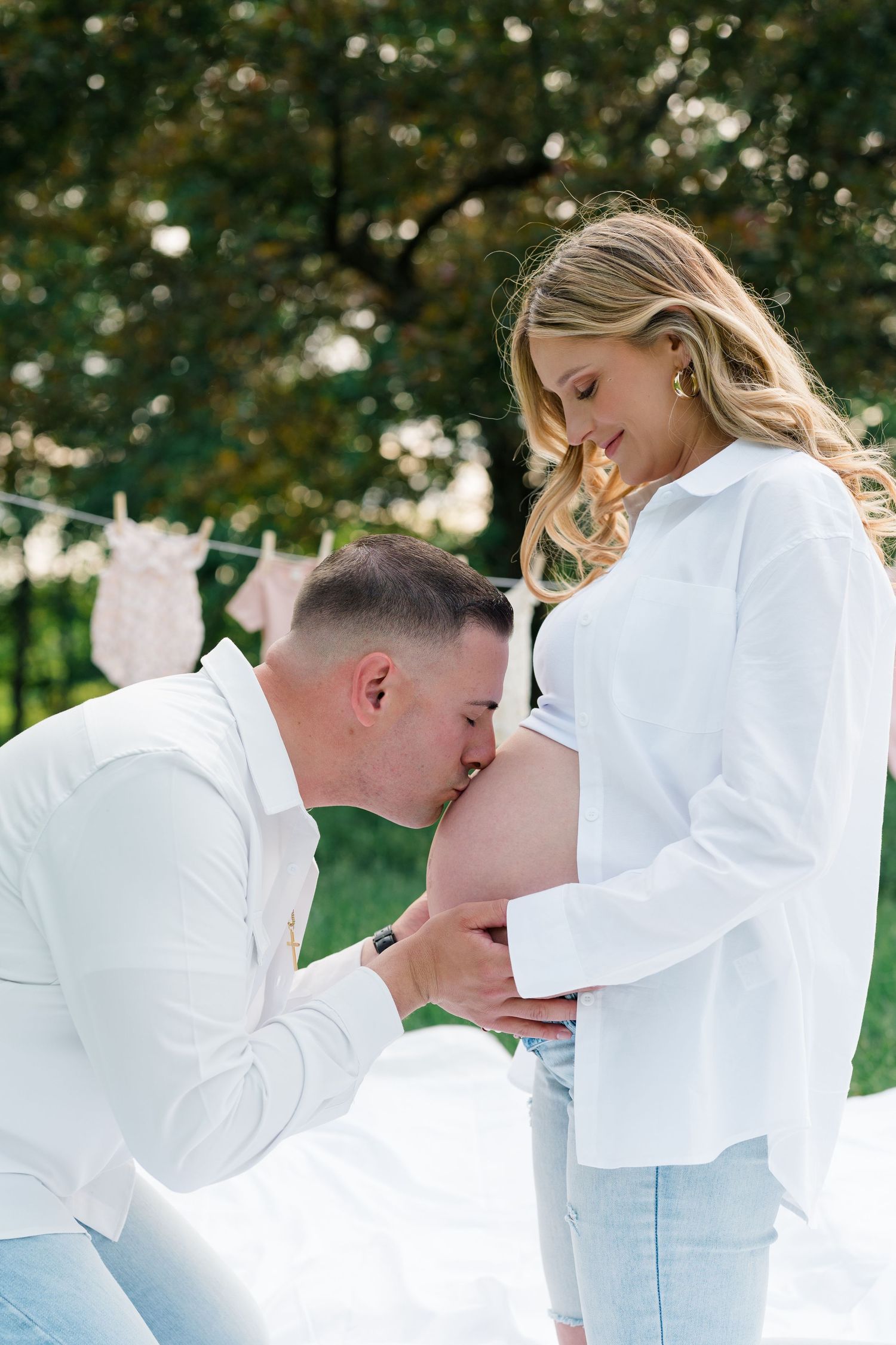 Romantic maternity photoshoot in a field with couple in white outfits embracing at sunset.