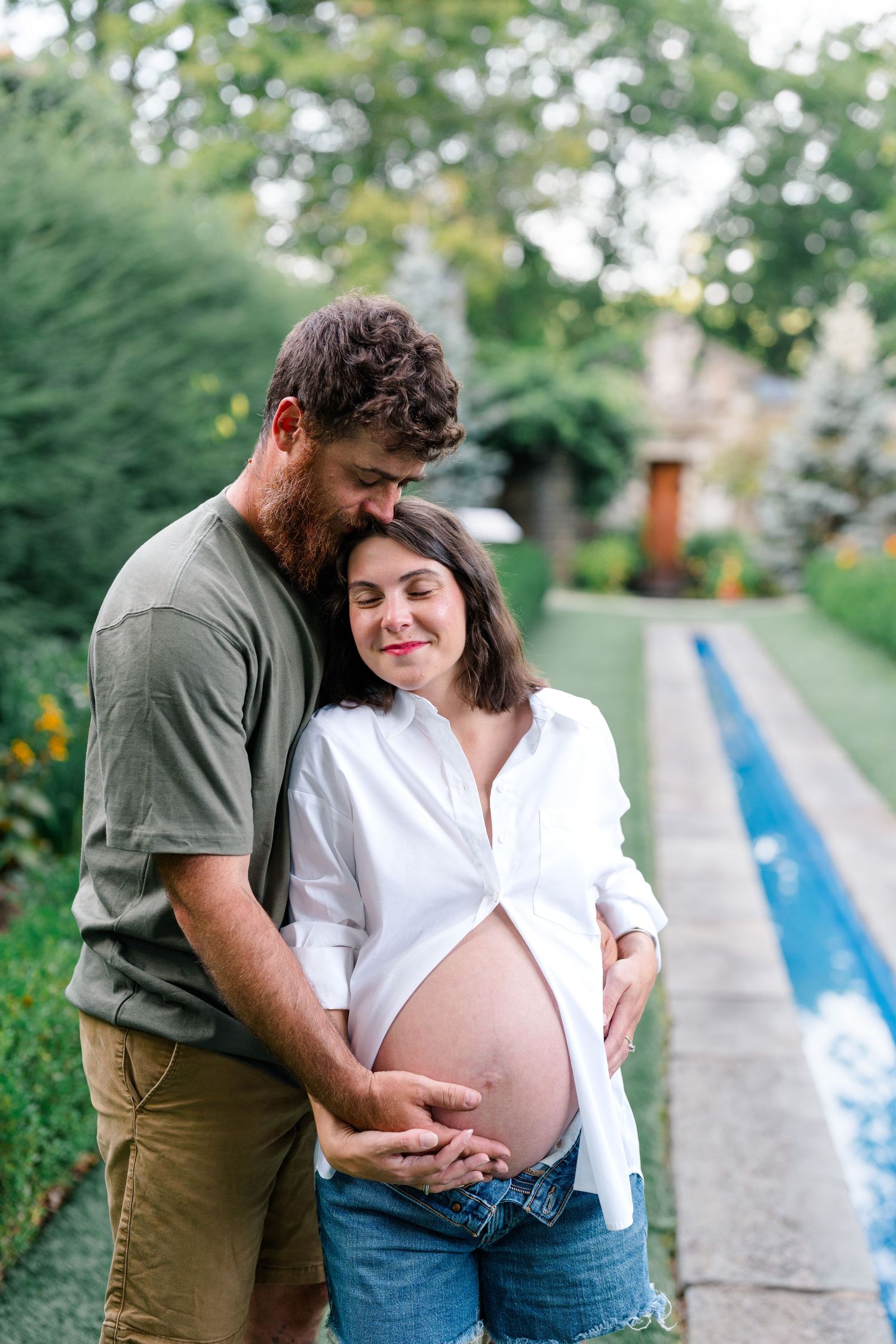 Expecting couple embracing outdoors, man holding pregnant woman's bare belly near a garden pool path.